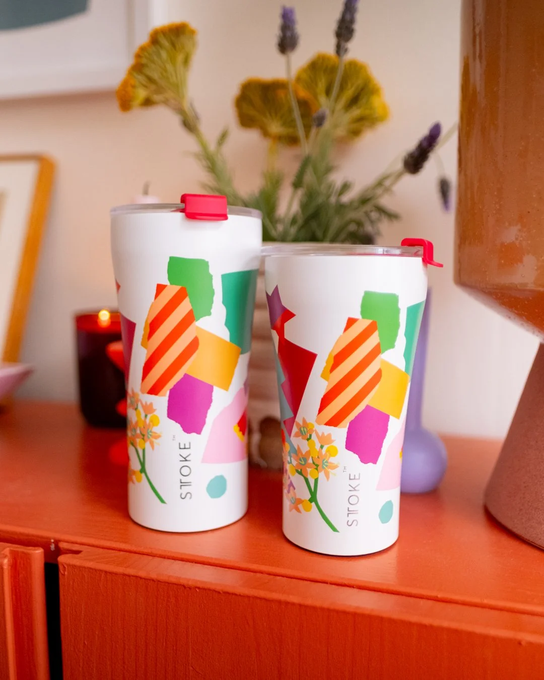 Two white STTOKE insulated tumblers in different sizes, both featuring a colourful collage-style print with geometric shapes and florals, styled on an orange sideboard with candles and dried flowers