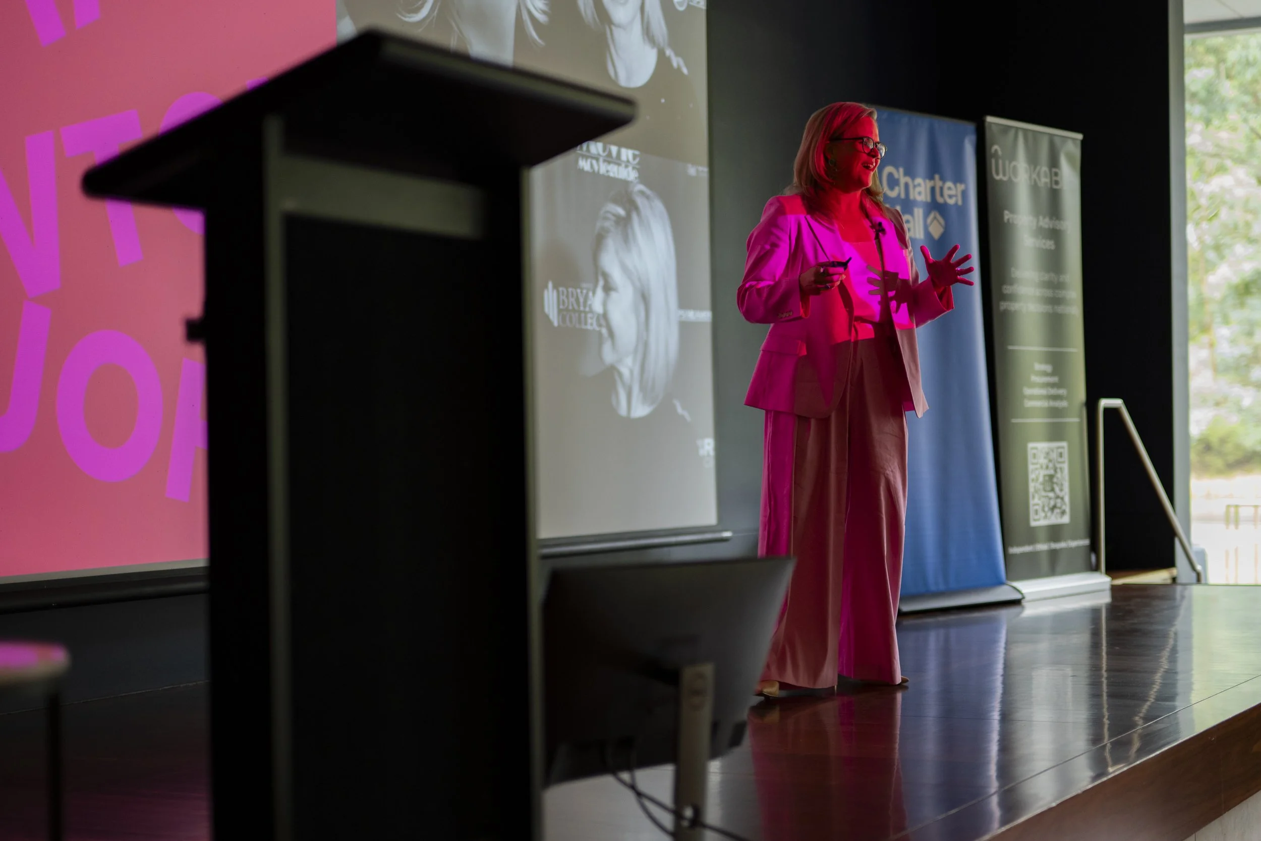 Taryn Brumfitt speaking on stage under pink stage lighting at a corporate event, with Charter Hall and Workable sponsor banners visible