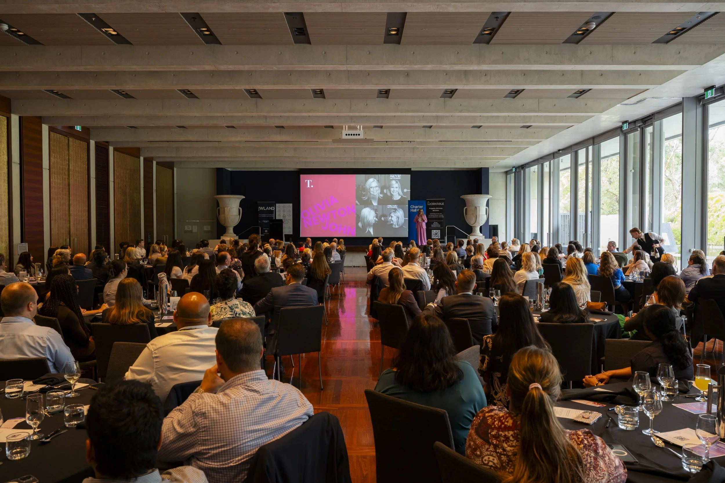 Wide shot of a full corporate luncheon audience seated at round tables, facing a speaker on stage at the National Gallery of Australia