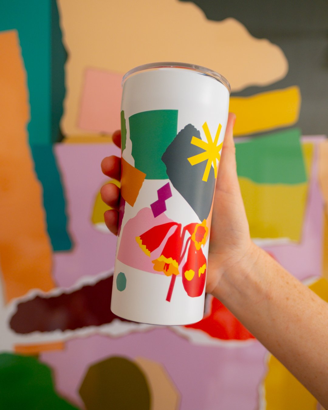 A hand holds up a white STTOKE tumbler featuring a bold collage-style print with geometric shapes and florals, set against a background of large torn coloured paper pieces in the style of Leah Bartholomew's artwork