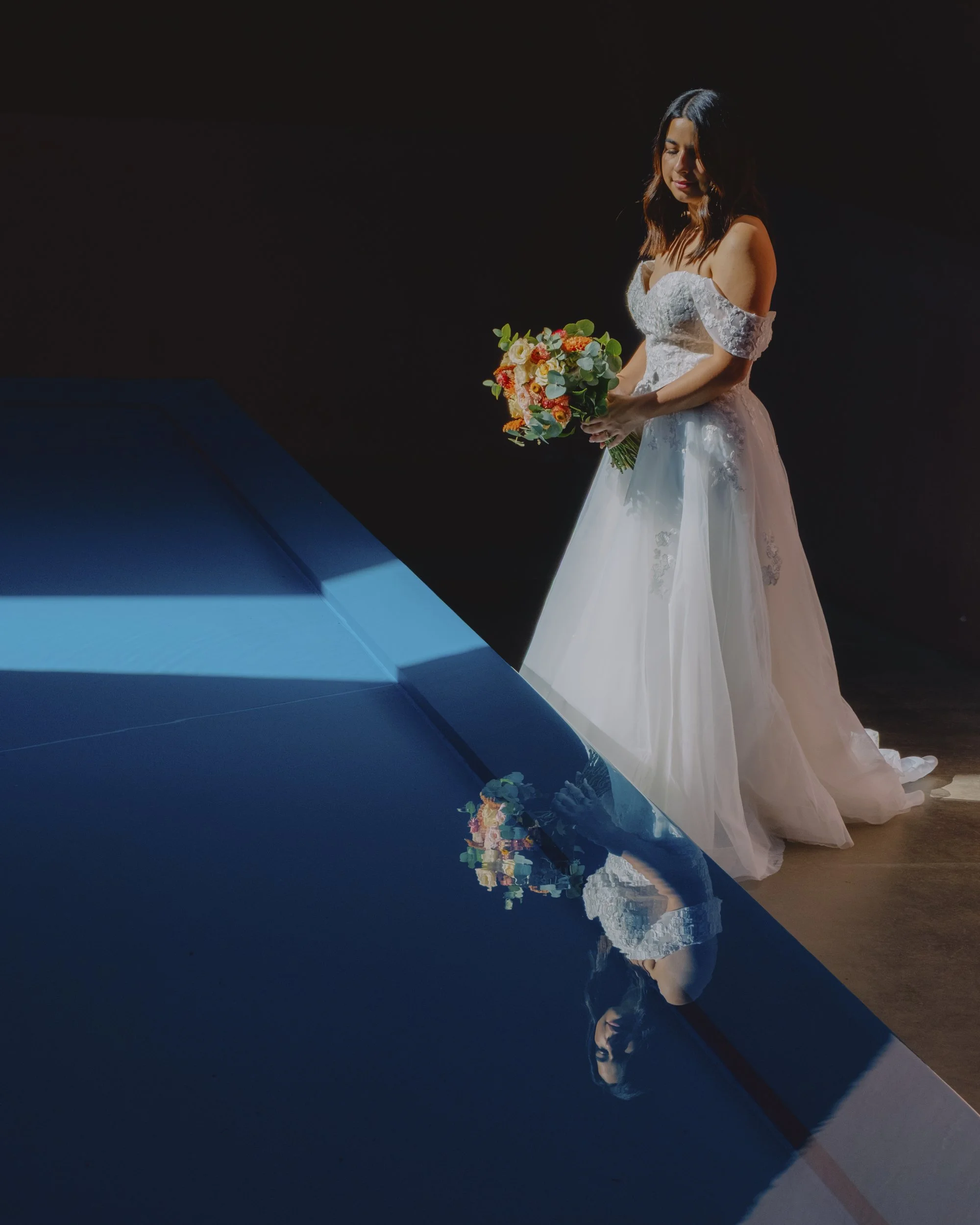 Bride standing at the edge of the reflecting pool inside the James Turrell Skyspace, Canberra, gown and bouquet reflected in the water, dramatic blue light