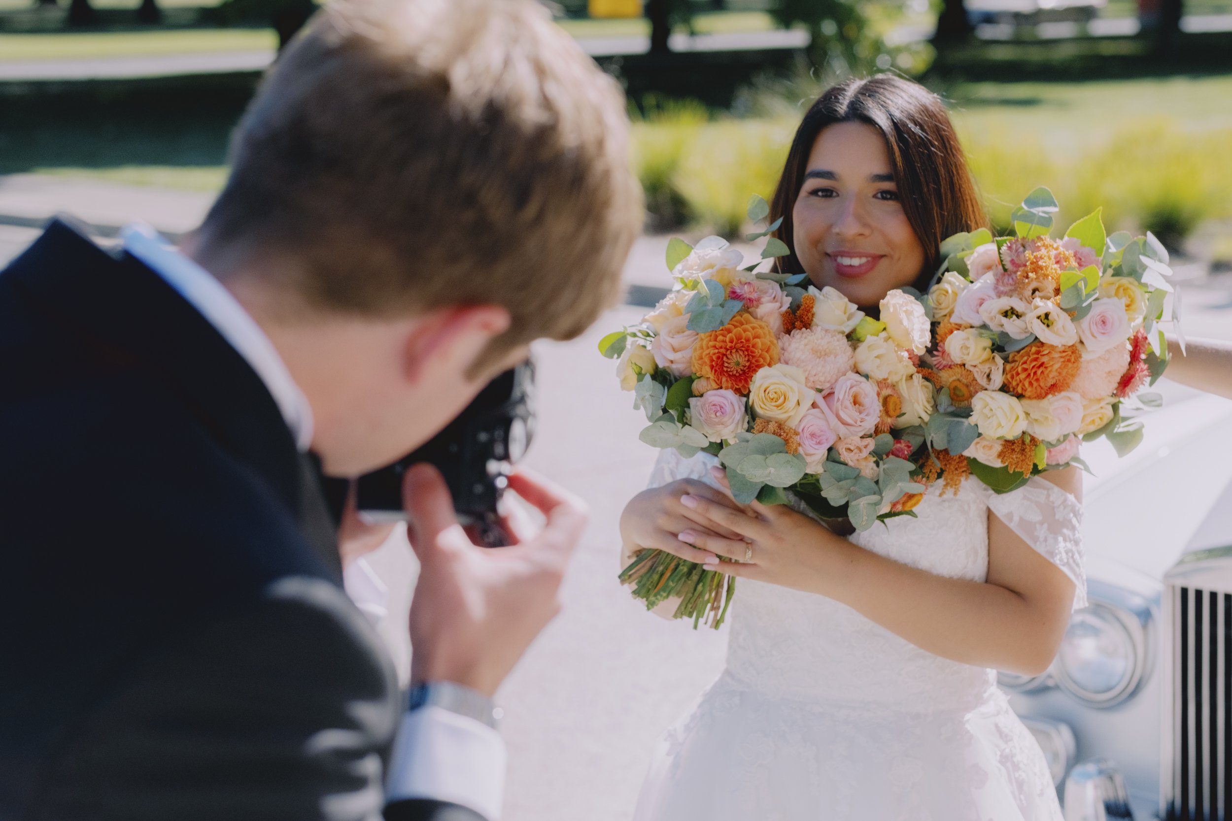 Groom photographing bride with a camera as she smiles over a large colourful bouquet, beside a vintage car