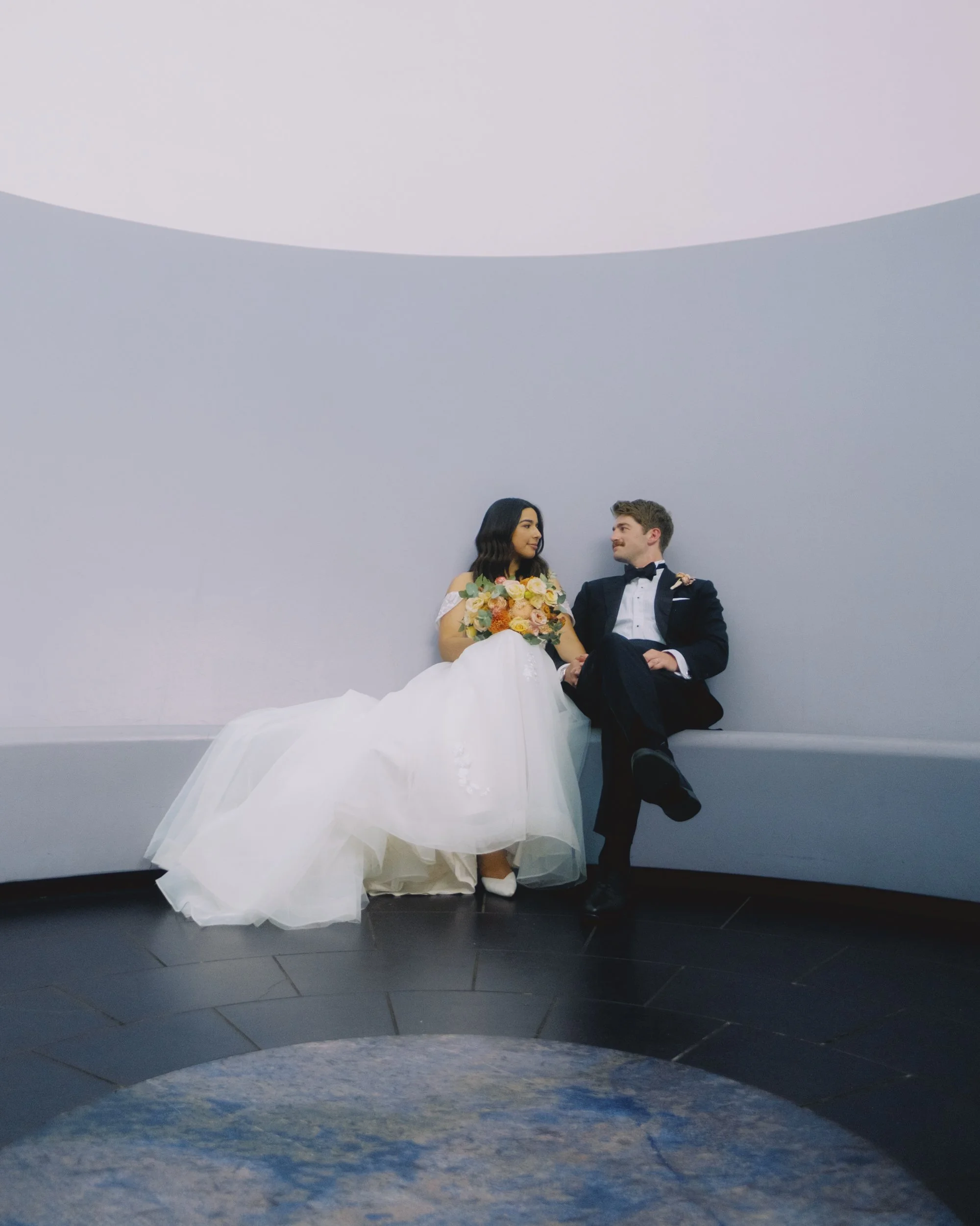 Bride and groom seated together on the curved bench inside the James Turrell Skyspace near the NGA, Canberra, soft diffused light filling the domed chamber