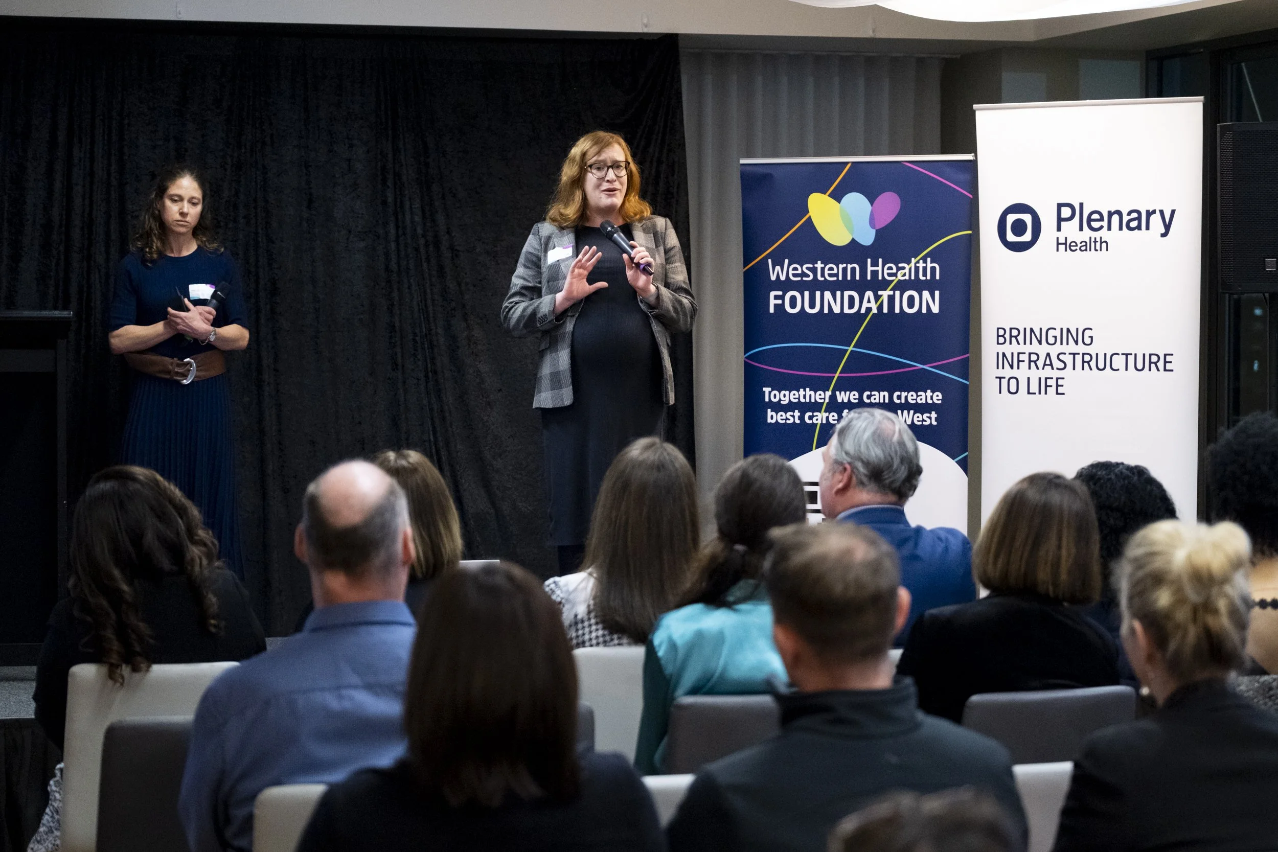 Speaker addressing a seated audience at a Western Health Foundation corporate event, with Western Health Foundation and Plenary Health banners on stage