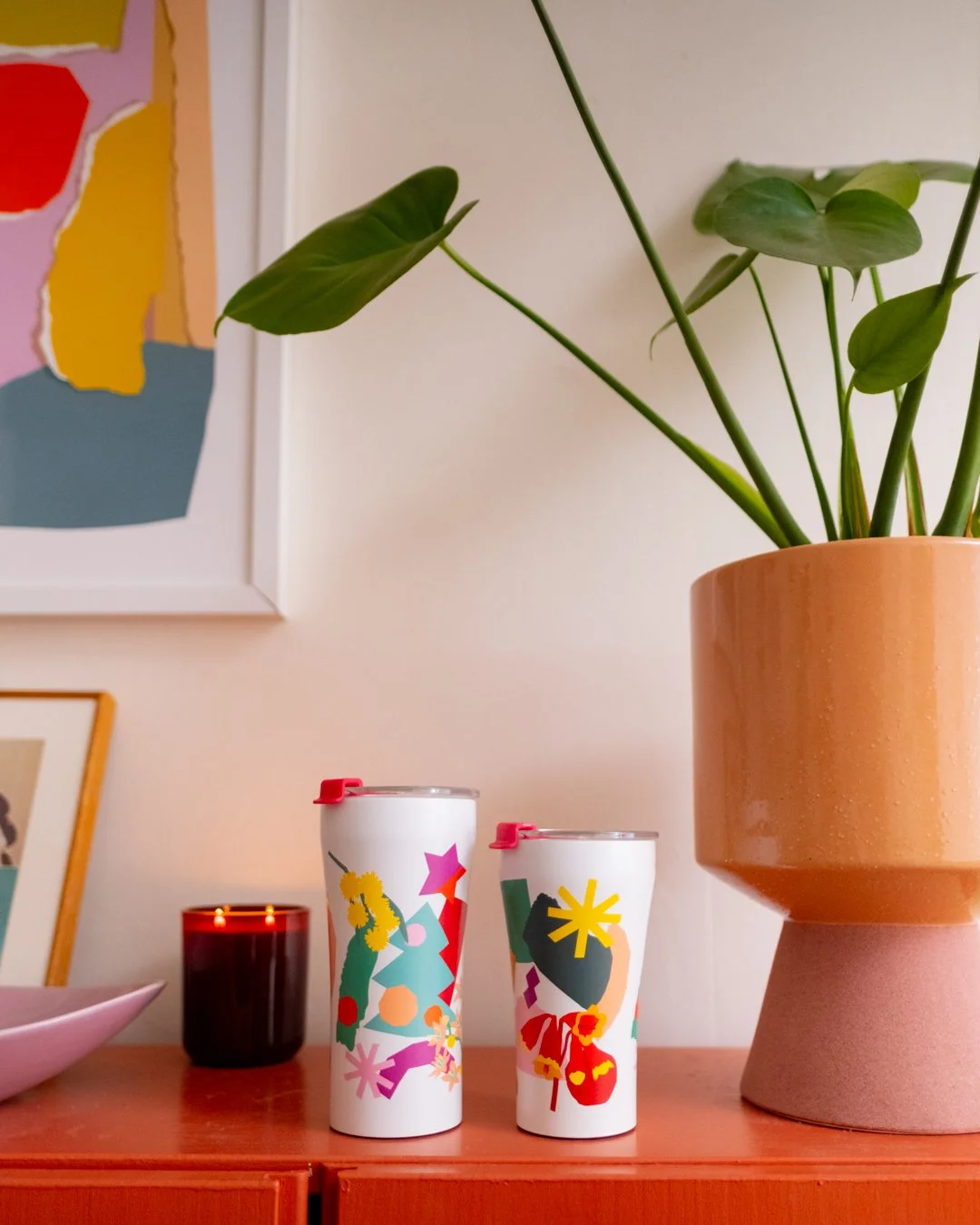 Two white STTOKE tumblers in different sizes with vibrant collage-style prints, displayed on an orange sideboard alongside a terracotta pot plant and abstract wall art
