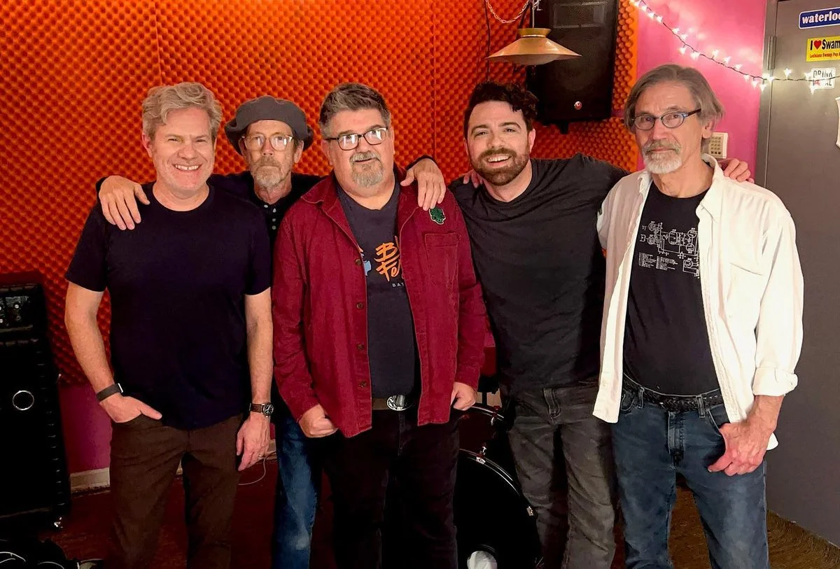 Five men standing together inside a room with red and pink foam wall panels, smiling and posing for a photo.
