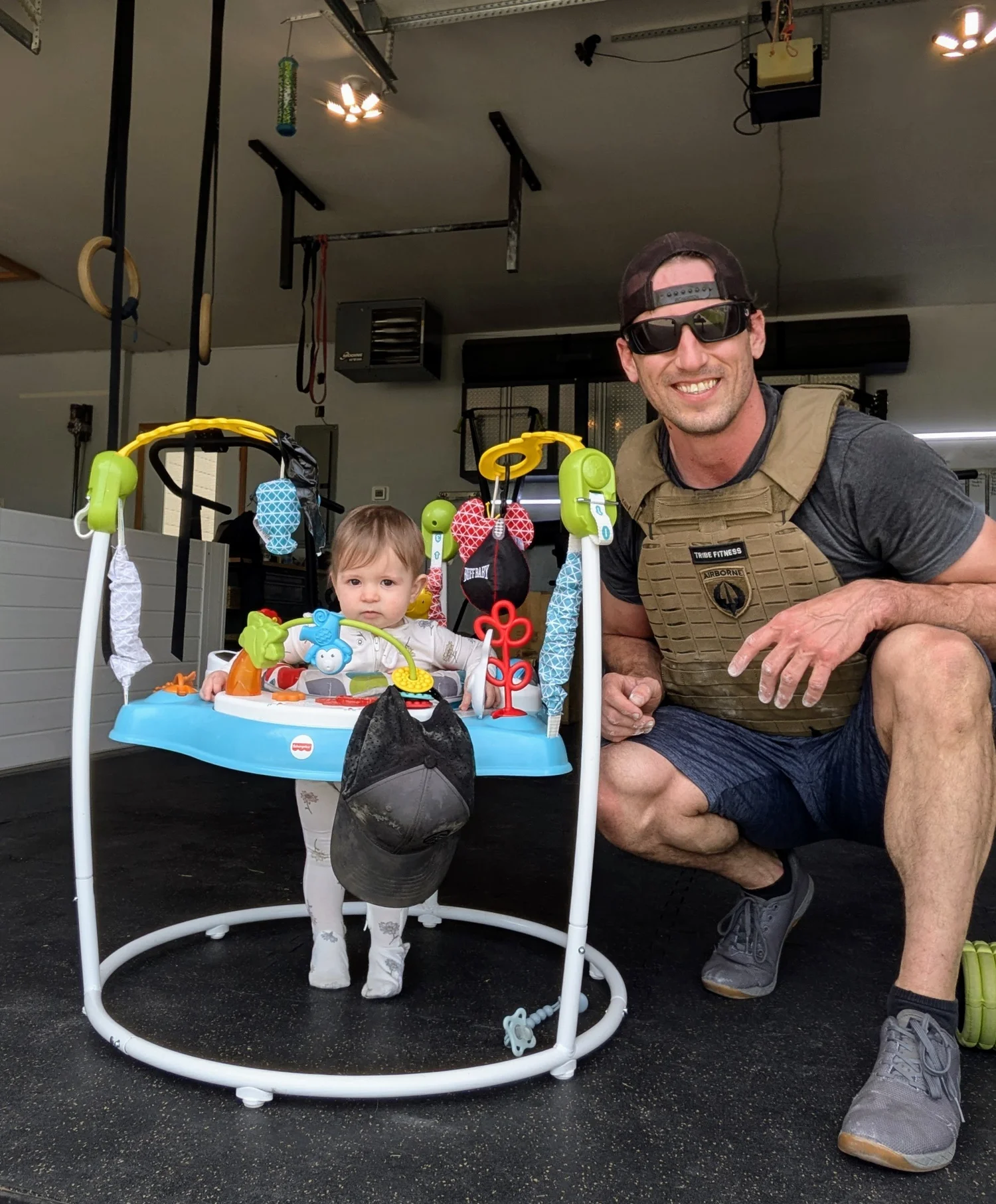 Coach Sam Moses in a weight vest at the Tribe Fitness garage studio in Mead, CO, next to his little one in a baby jumper