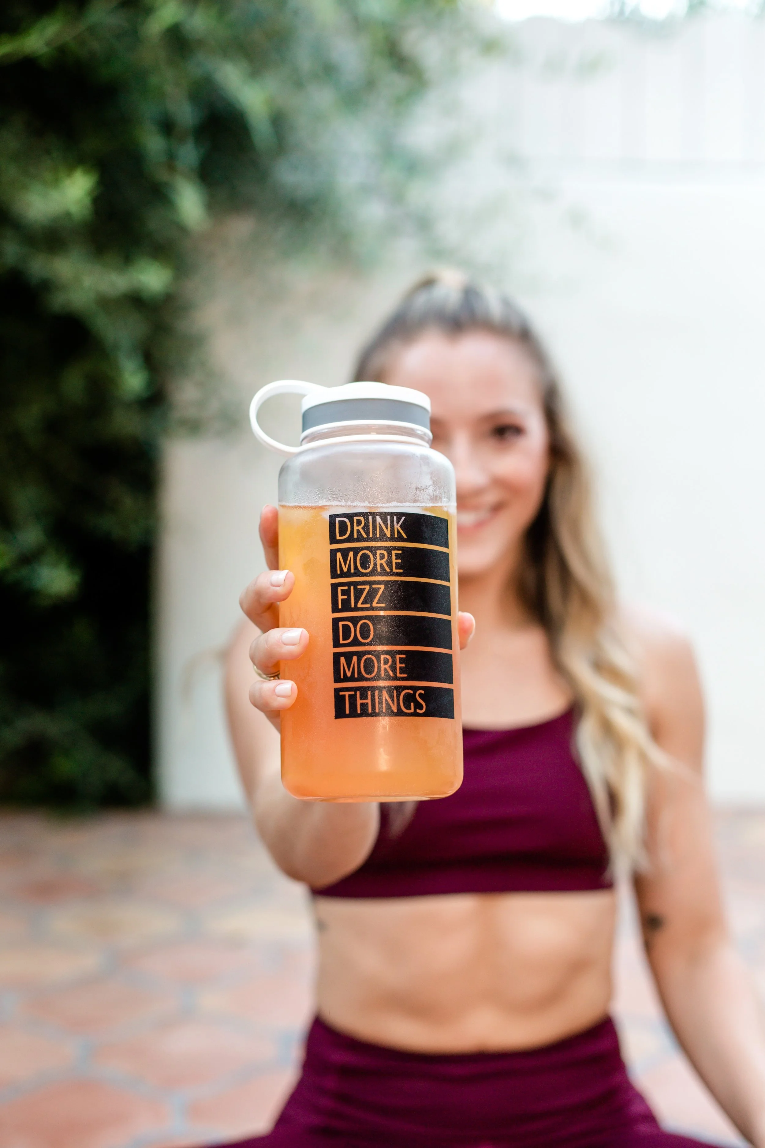 Close-up of a fitness drink bottle labeled ‘Drink More Fizz, Do More Things’ held by a smiling woman in burgundy workout wear during a branding session in Pasadena