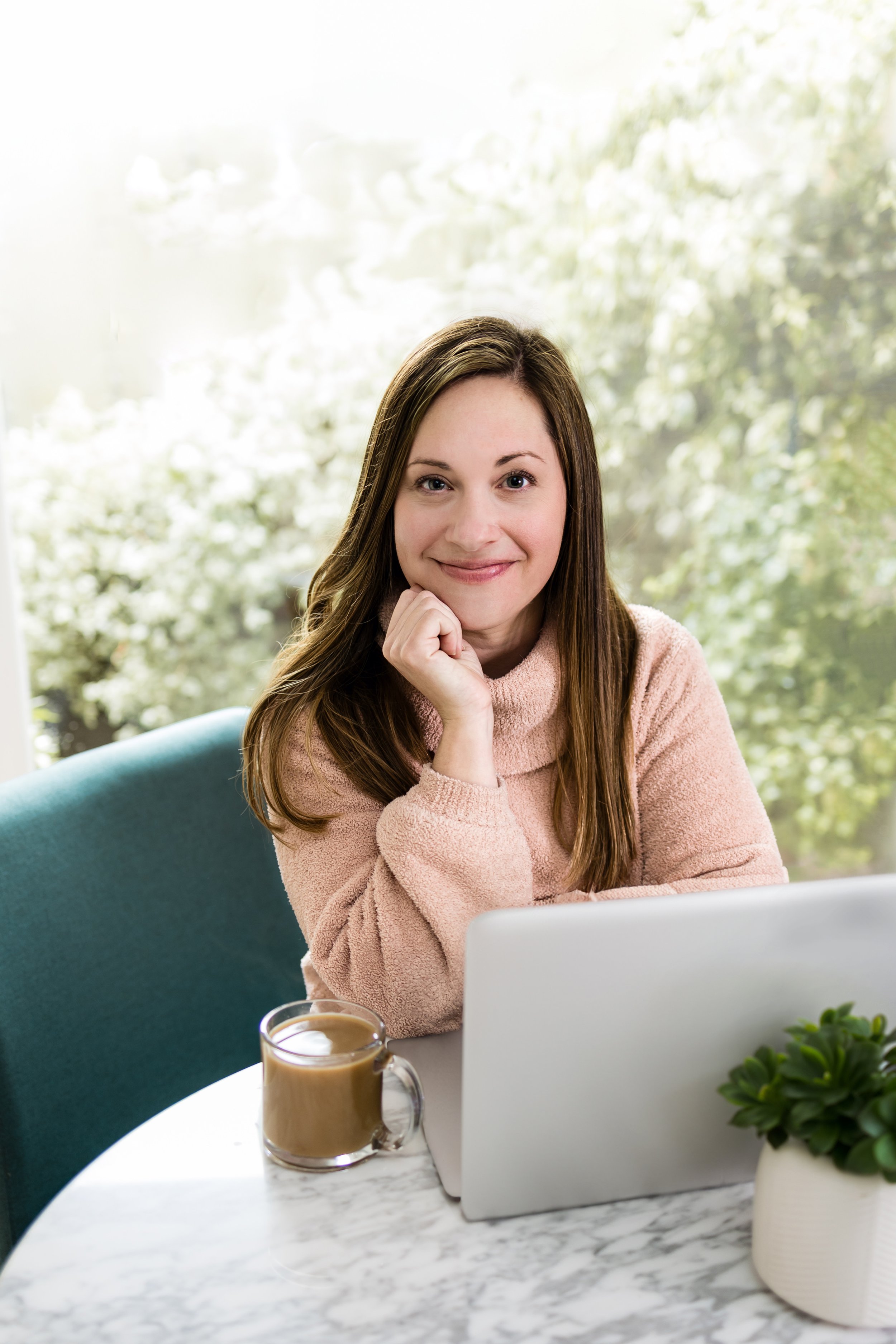 Personal branding photo of a woman entrepreneur smiling at a laptop with a coffee mug, seated at a marble table near a window with greenery in the background