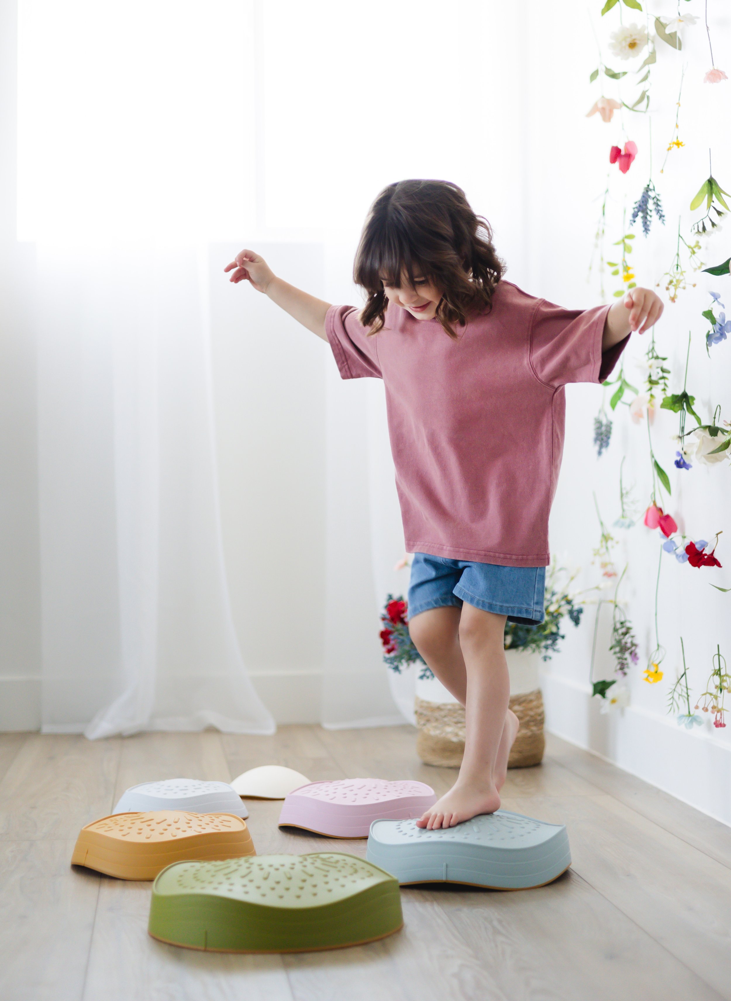 A young girl with wavy brown hair, wearing a mauve T-shirt and denim shorts, balancing on a light gray balance board in a bright room with white walls. There are additional colorful balance boards on the wooden floor and a backdrop decorated with hanging artificial flowers.
