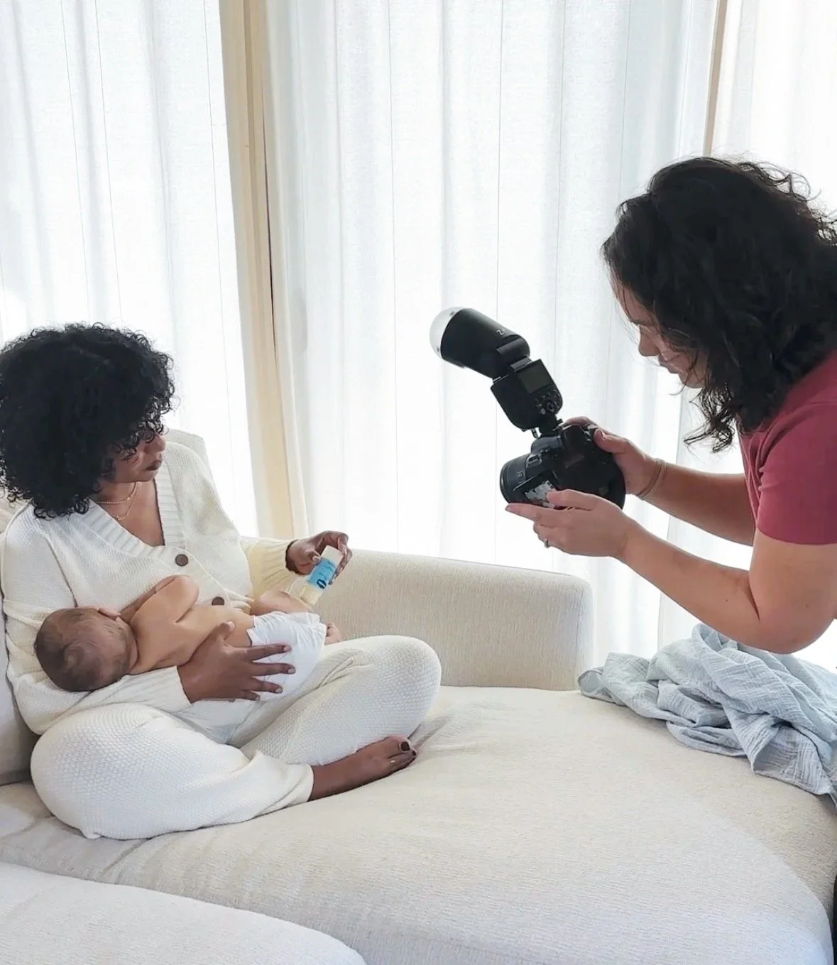 Woman breastfeeding a baby while another woman takes a photograph in a bright room with white curtains.