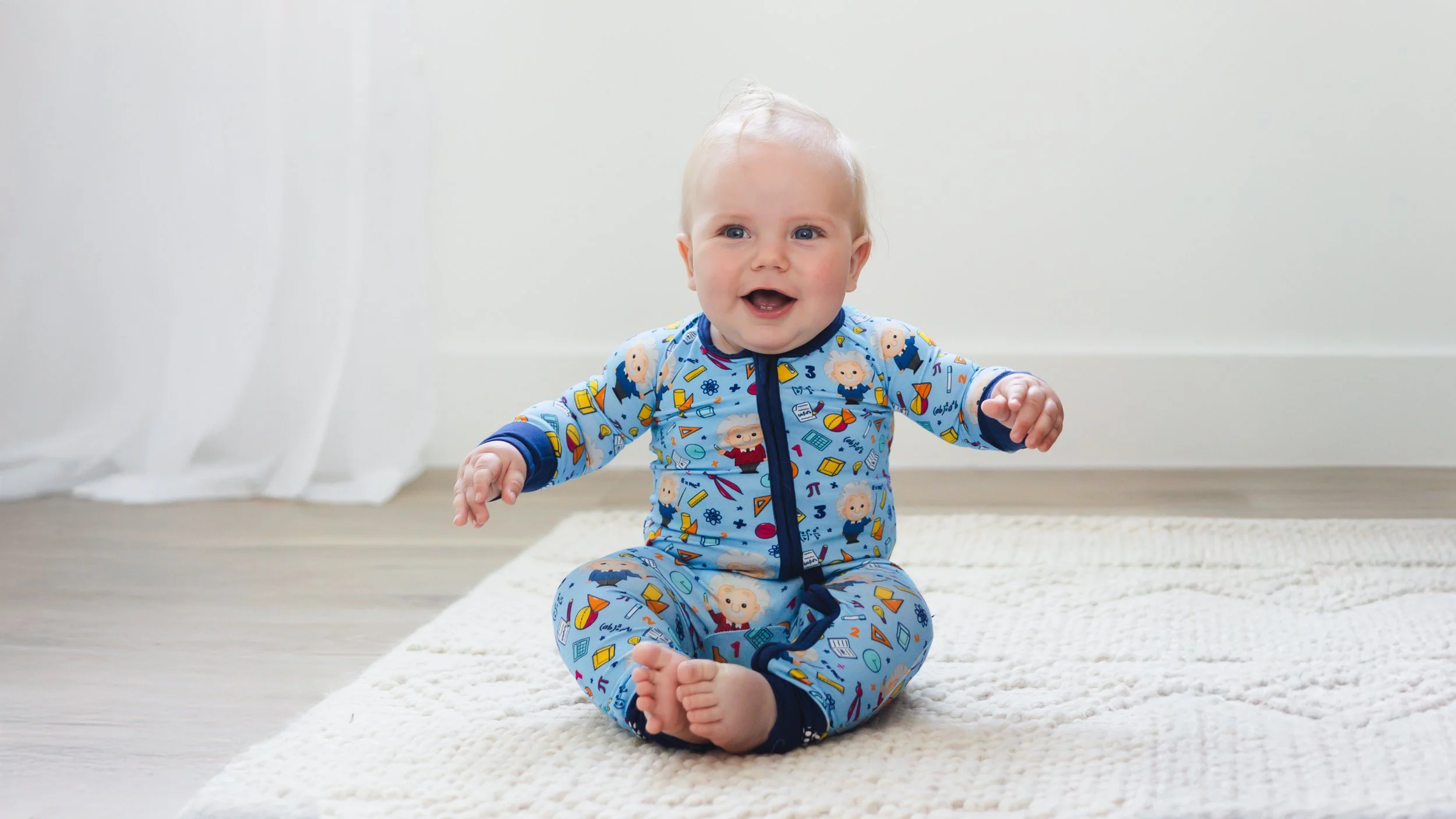A smiling baby sitting on a white rug at home, wearing a blue onesie with colorful science-themed and cartoon faces, with arms outstretched and a happy expression.