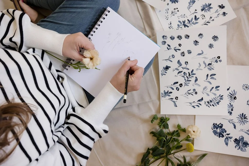 Flat lay of artist sketching botanical flowers in a notebook surrounded by floral illustrations and greenery during a creative branding session