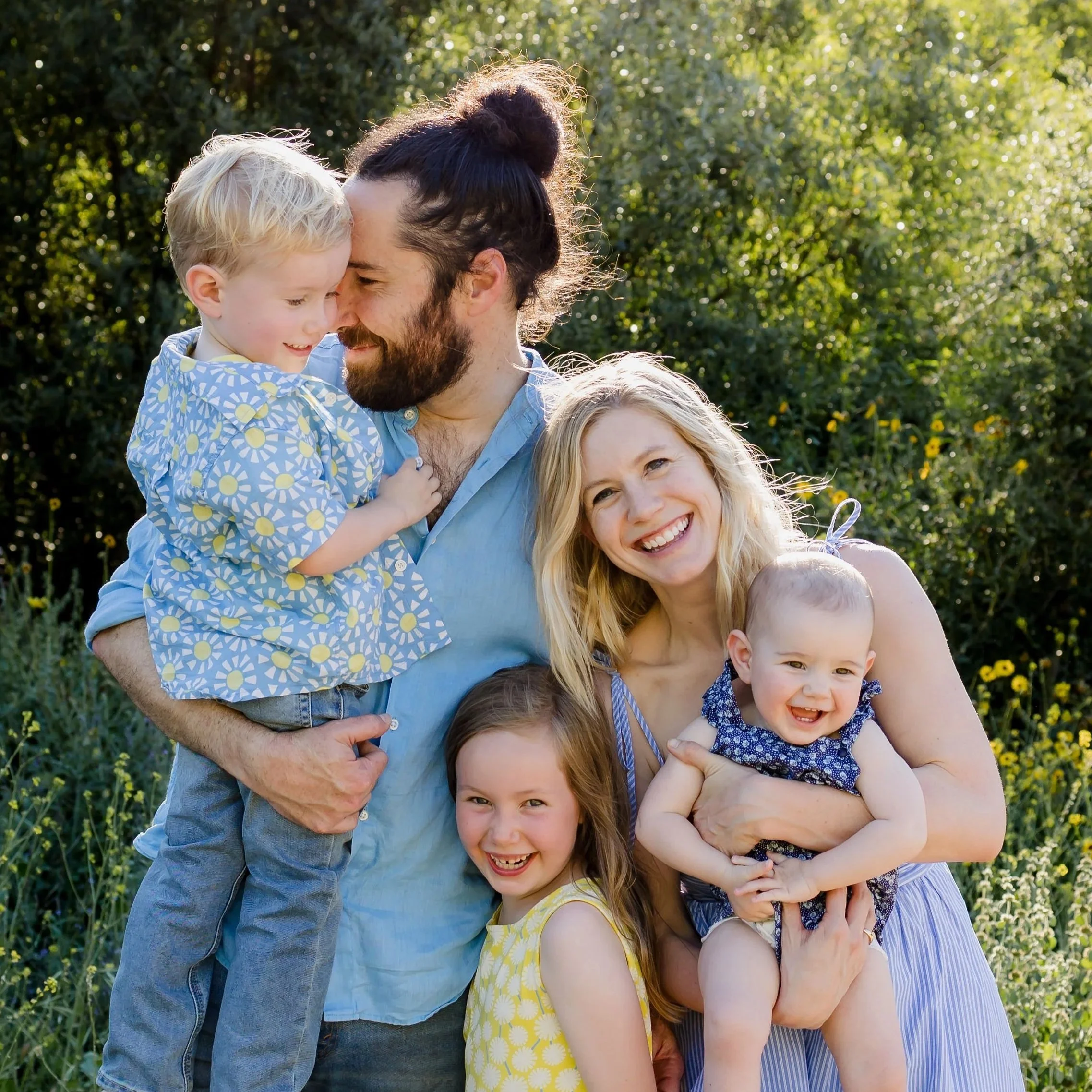 A young family wearing blue and yellow, smiling, standing outdoors in front of yellow-flowered bushes.