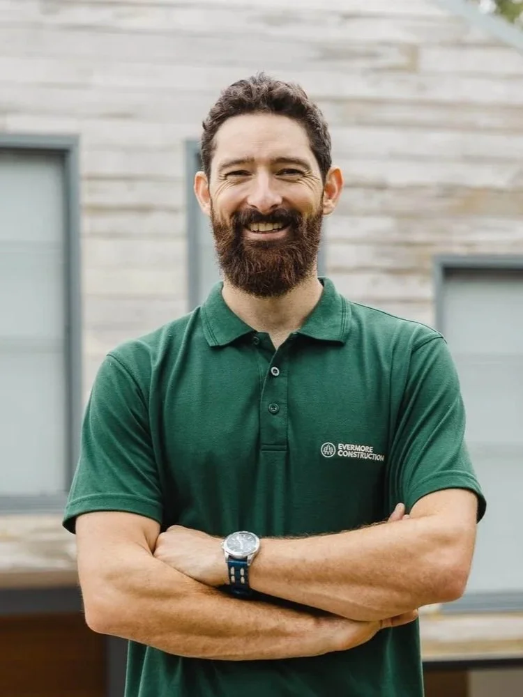Man with a dark beard, wearing a green polo shirt with a company logo, smiling with arms crossed, standing outside in front of a building with grey wooden siding and windows.