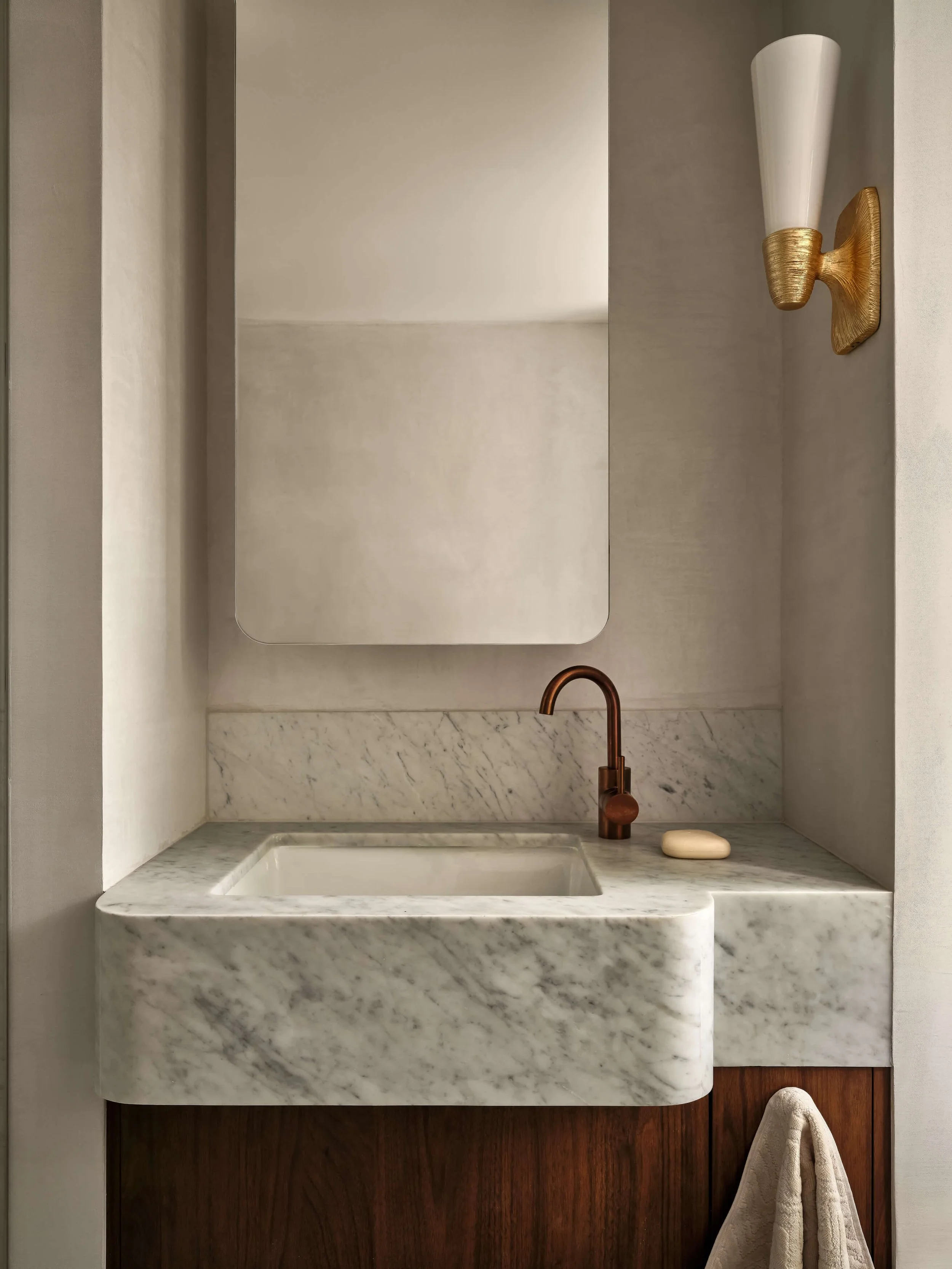 A minimalist bathroom vanity with a marble sink, copper faucet, a mirror, a wall sconce, and a towel hanging below.