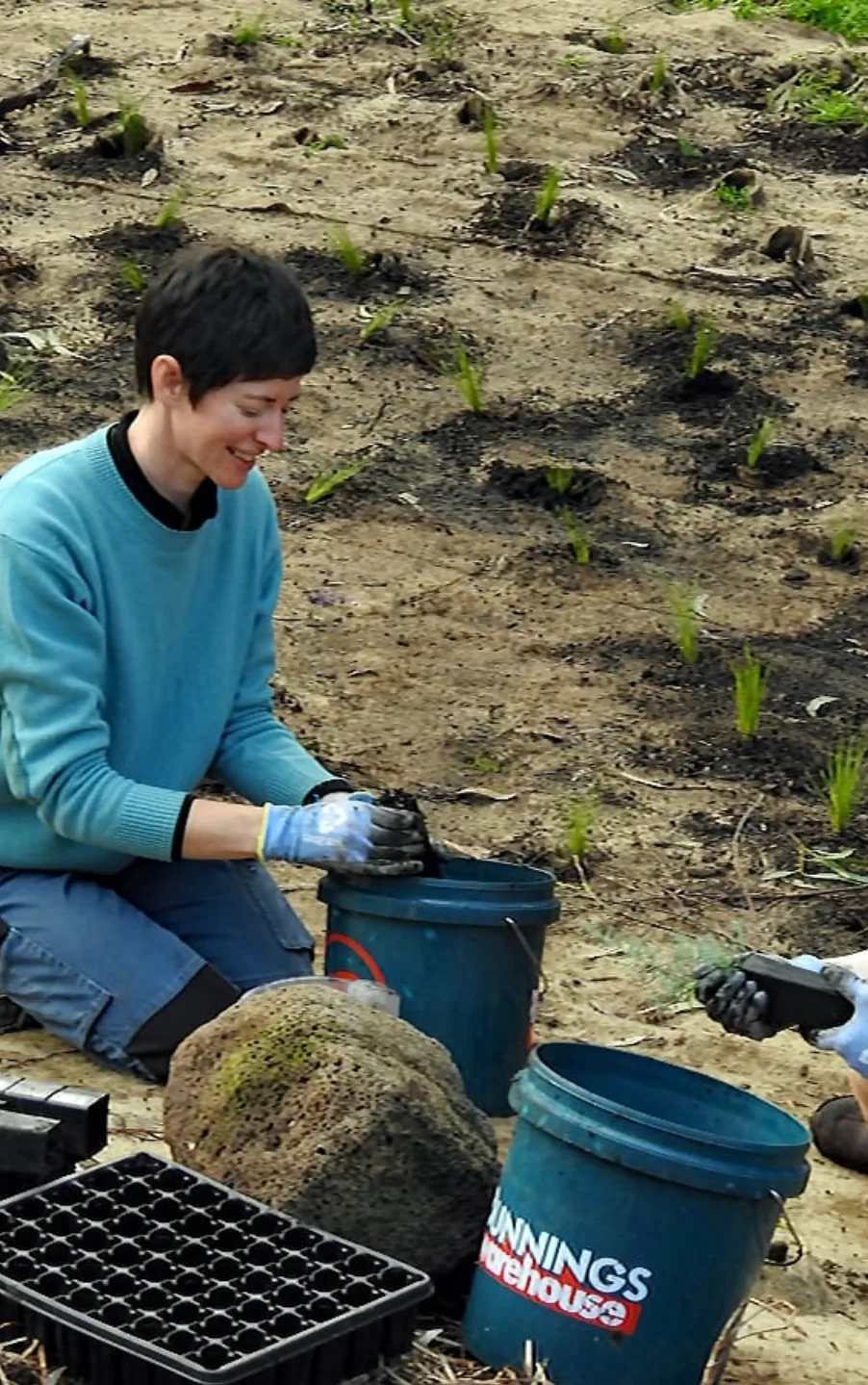 A woman with short brown hair smiling as she plants a seedling