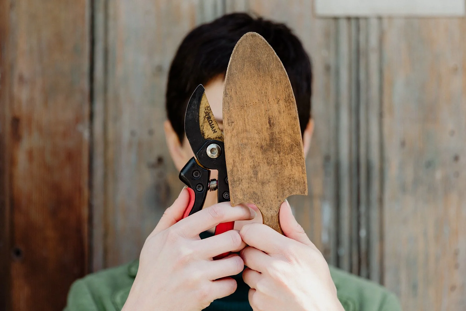 A white woman holding up secateurs and a wooden trowel in front of her face