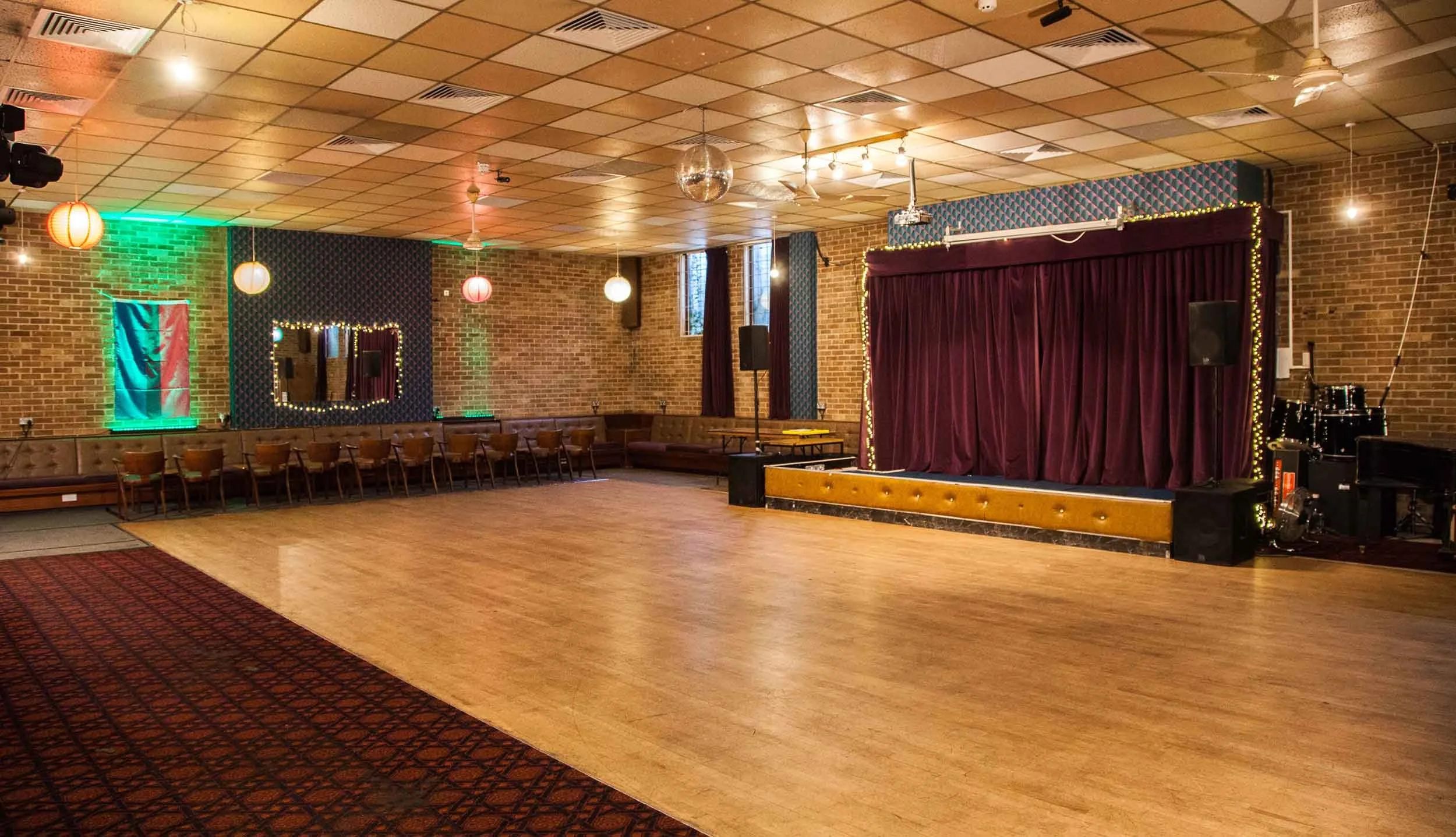 Wide hall with a wooden dance floor, small stage with purple curtains, brick walls, hanging lanterns, and bench seating along the sides.
