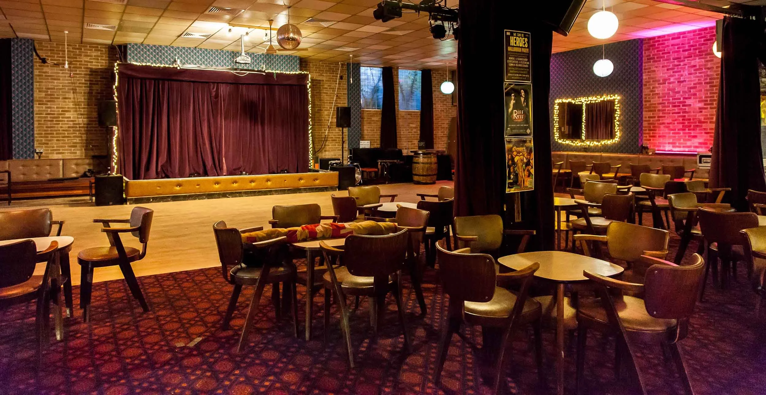 Tables and chairs on patterned carpet facing a wooden dance floor and small stage with purple curtains, with brick walls and hanging globe lights