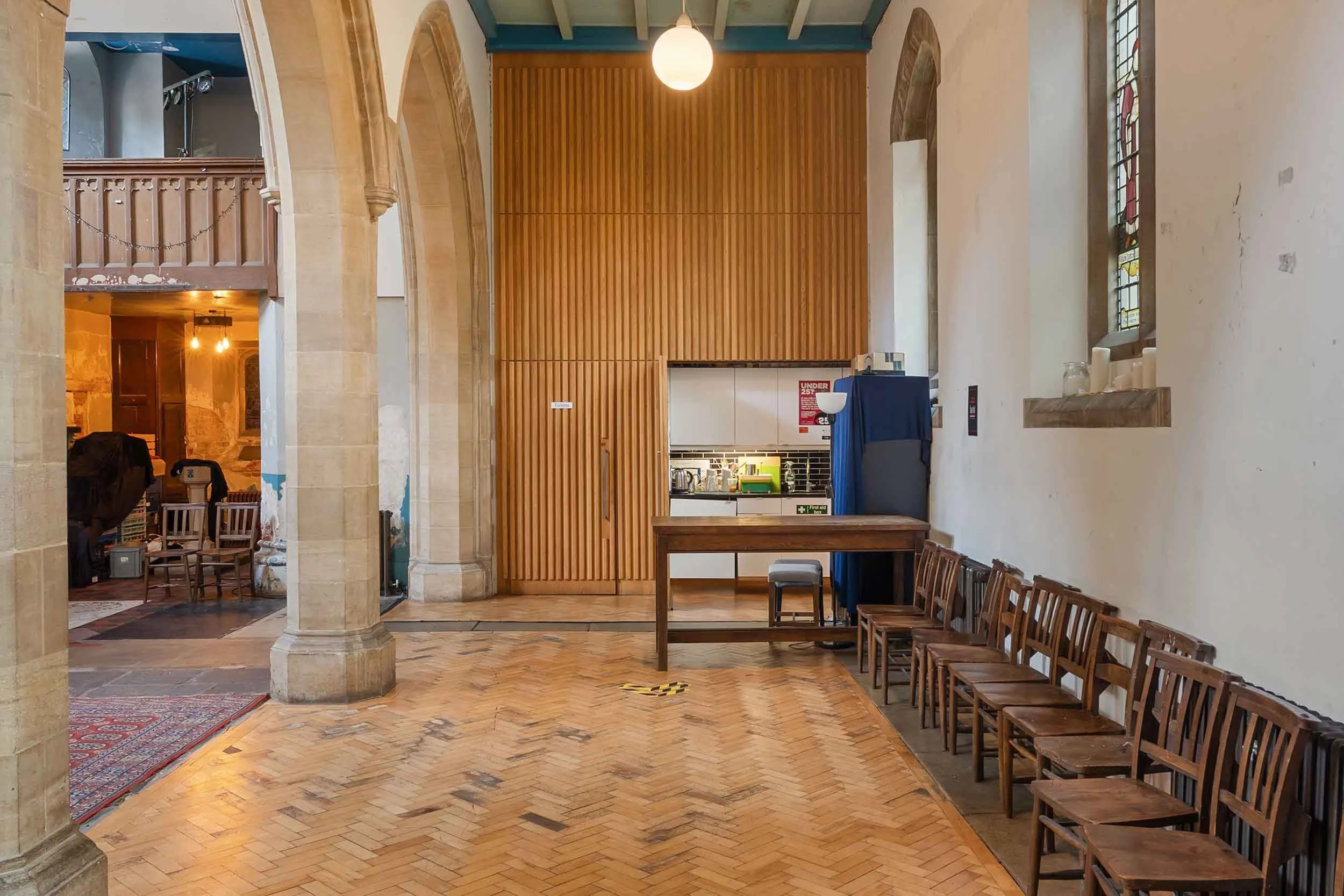 Herringbone floor with a row of wooden chairs, a long table, and a kitchenette set into a wood-panelled wall.