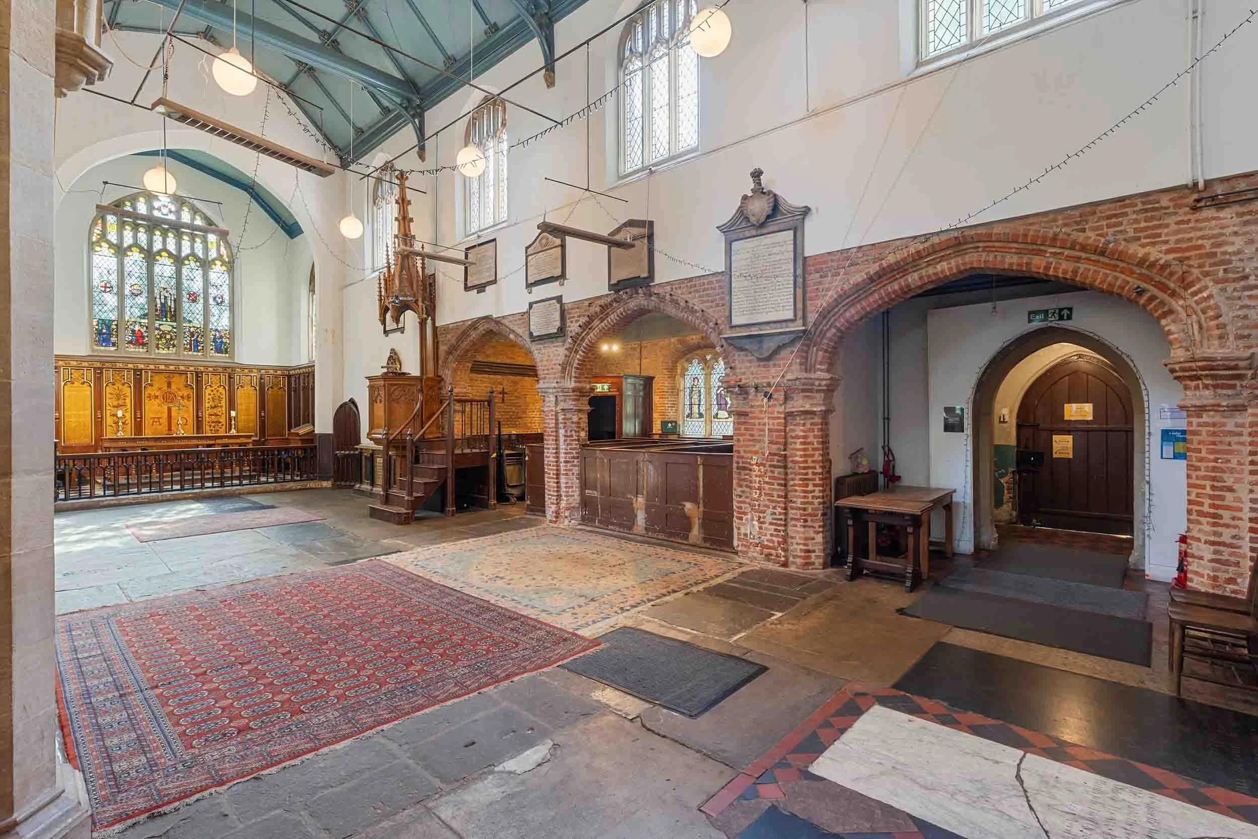 Wide interior with arched brick openings, stone floor and rugs, and stained-glass window at the far end.