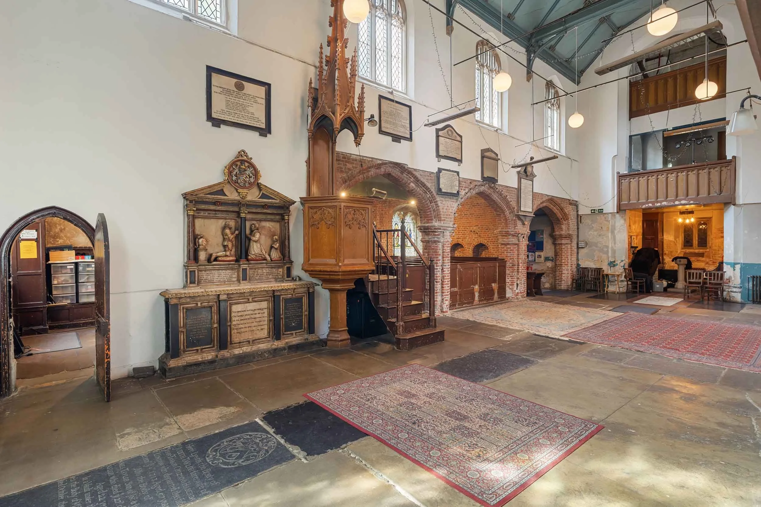 Wide hall with stone floor, patterned rugs, carved wooden pulpit, and arched brick openings under hanging globe lights.