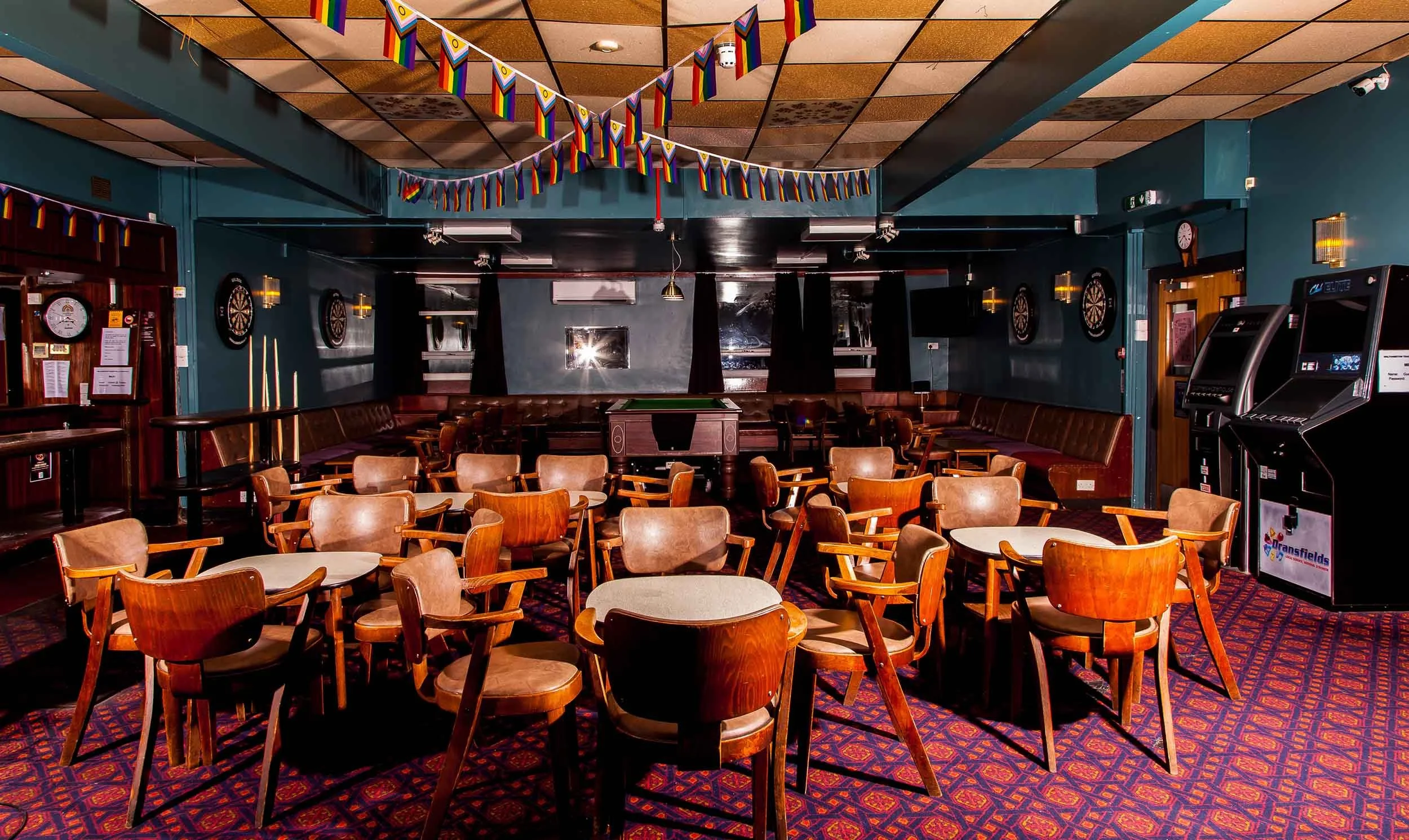 Hall with wooden chairs and round tables on patterned carpet, rainbow bunting overhead, darts boards on the walls, and arcade machines to one side.