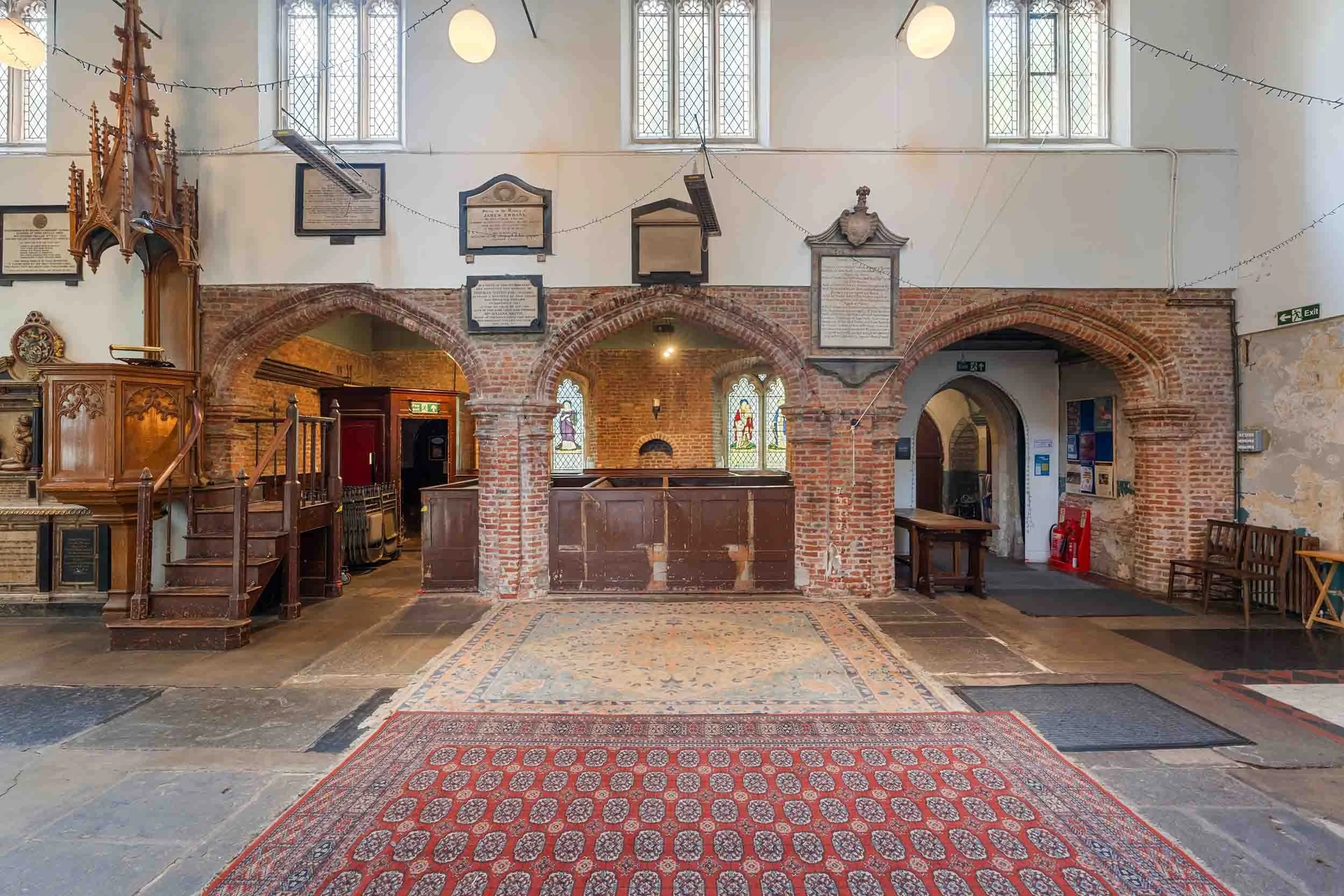 Brick arches and stone floor with patterned rugs, wooden pulpit, and memorial plaques beneath tall windows.