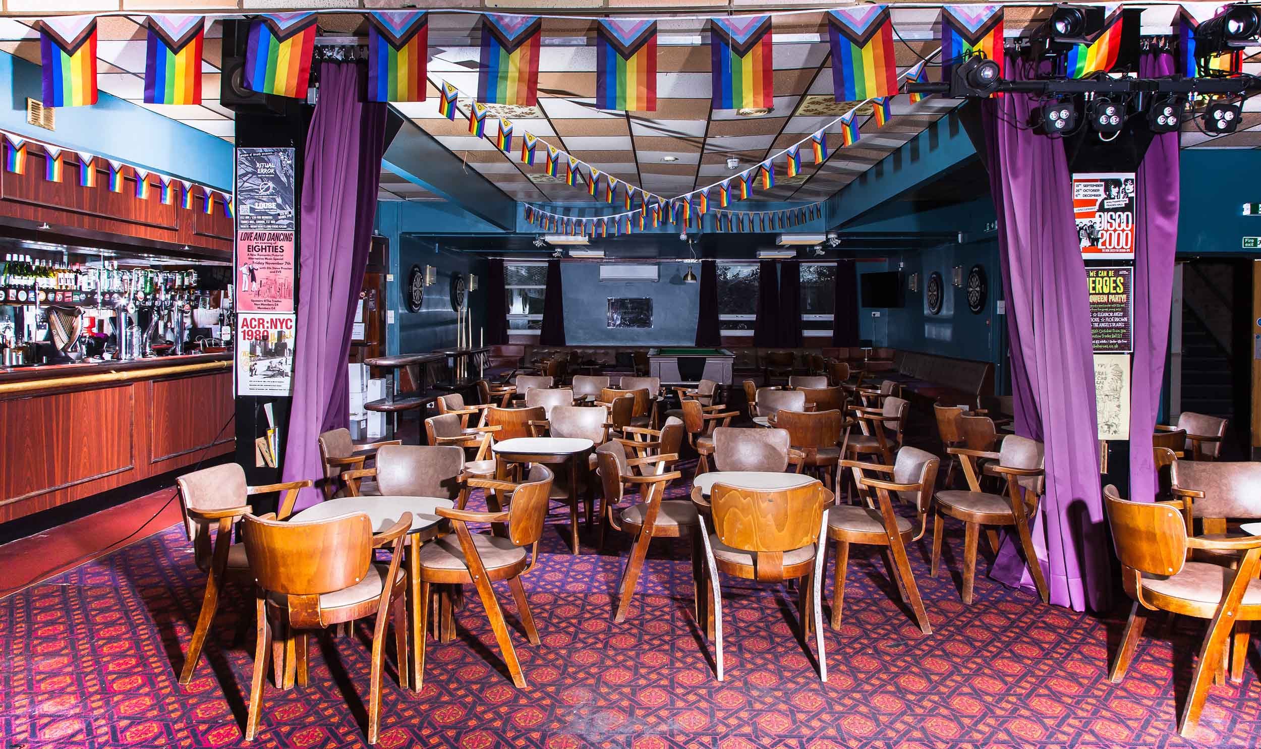 Bar and seating area with wooden chairs and tables, patterned carpet, purple curtains, and rainbow bunting hanging from the ceiling.