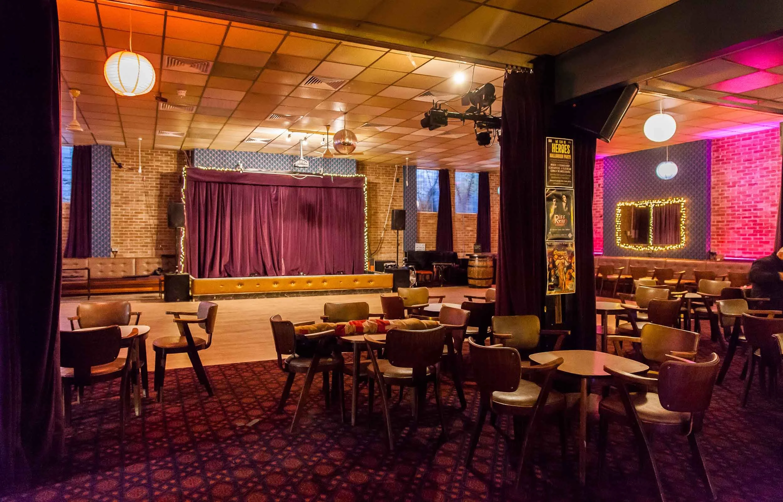 Seating area leading to a wooden dance floor and small stage with purple curtains, with hanging paper lanterns and brick walls.