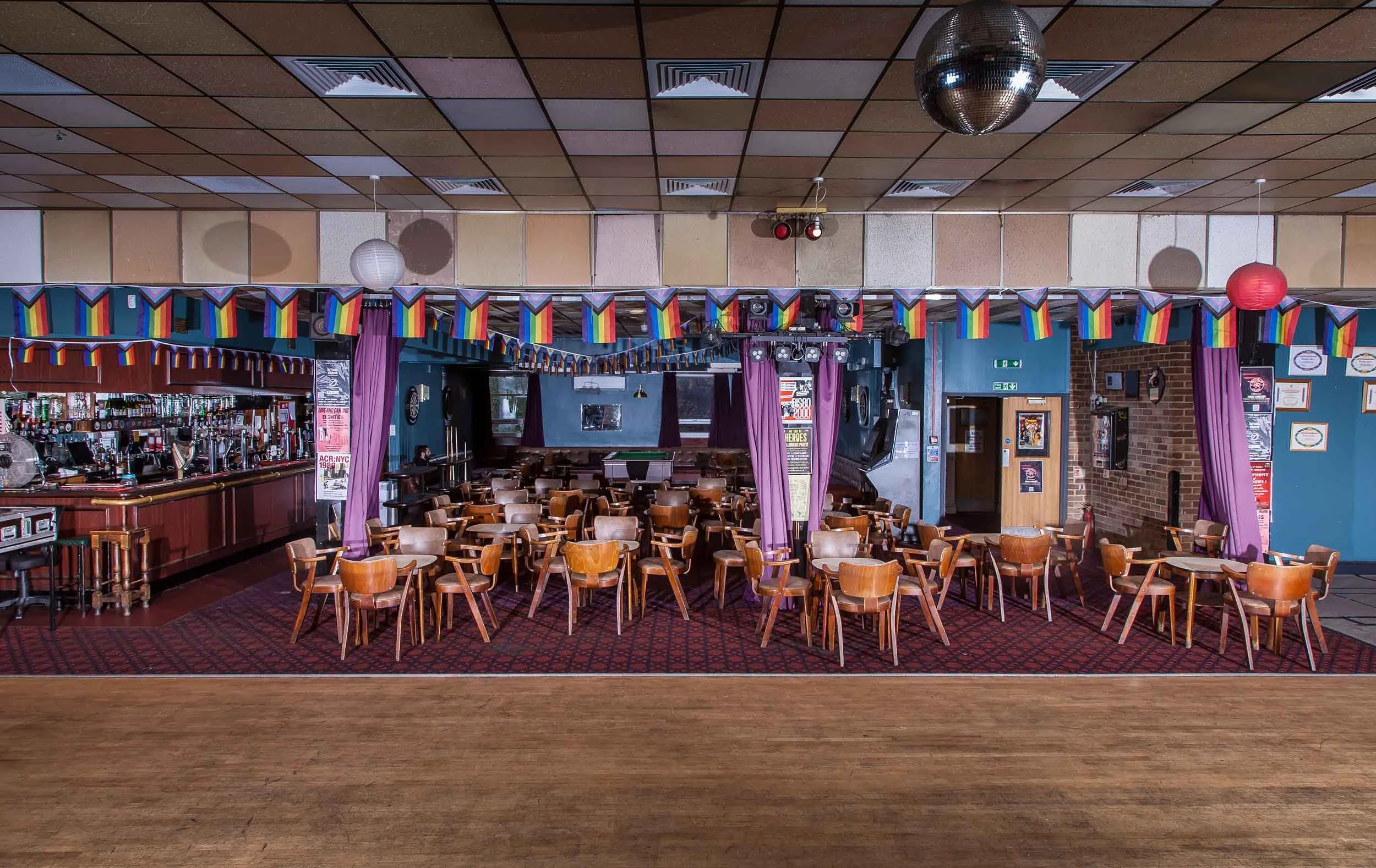 Wide hall with wooden dance floor, tables and chairs, long bar to the left, disco ball, and rainbow bunting across the ceiling.
