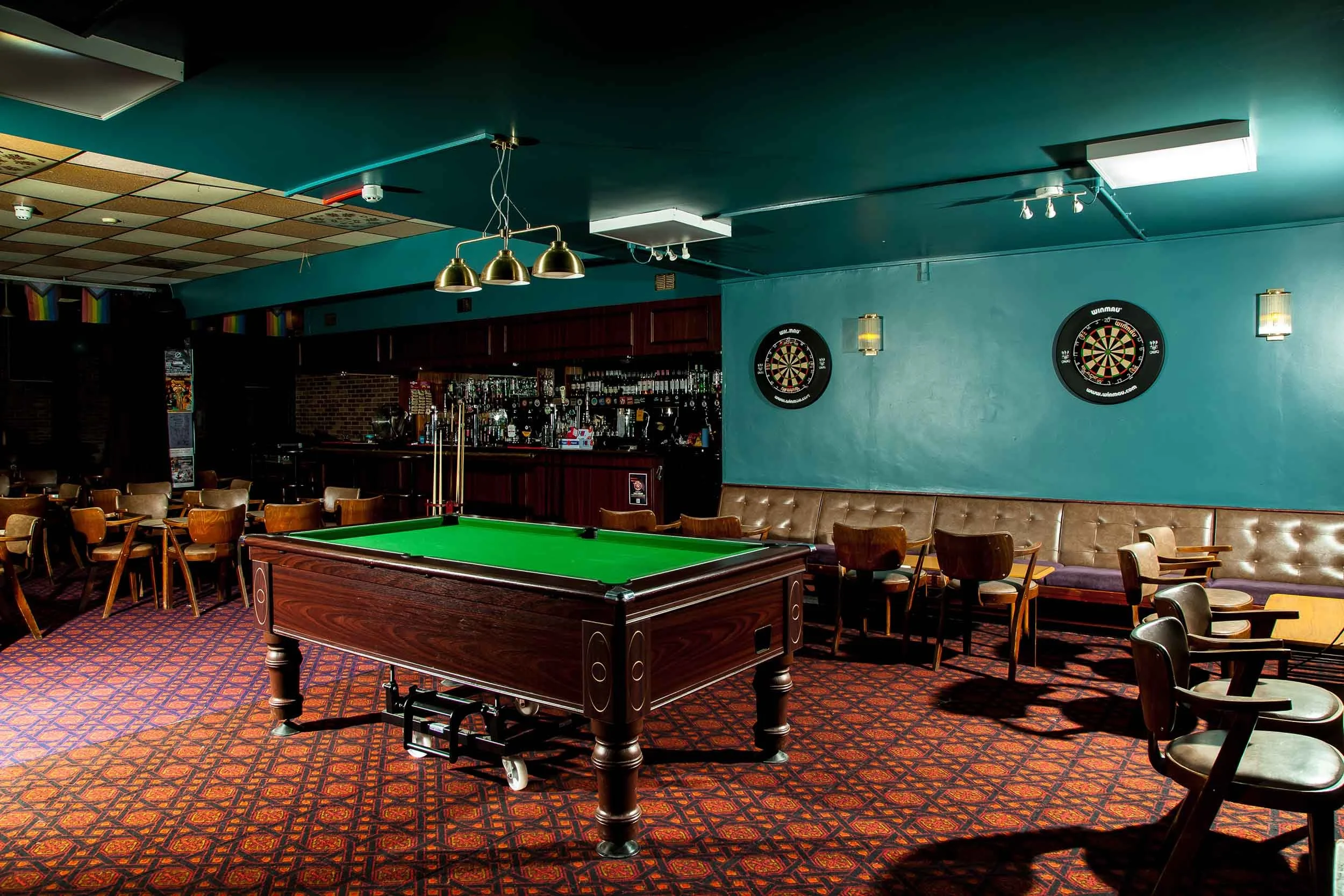 Snooker table on patterned carpet with bench seating along a blue wall, dartboards above, and a bar area in the background.