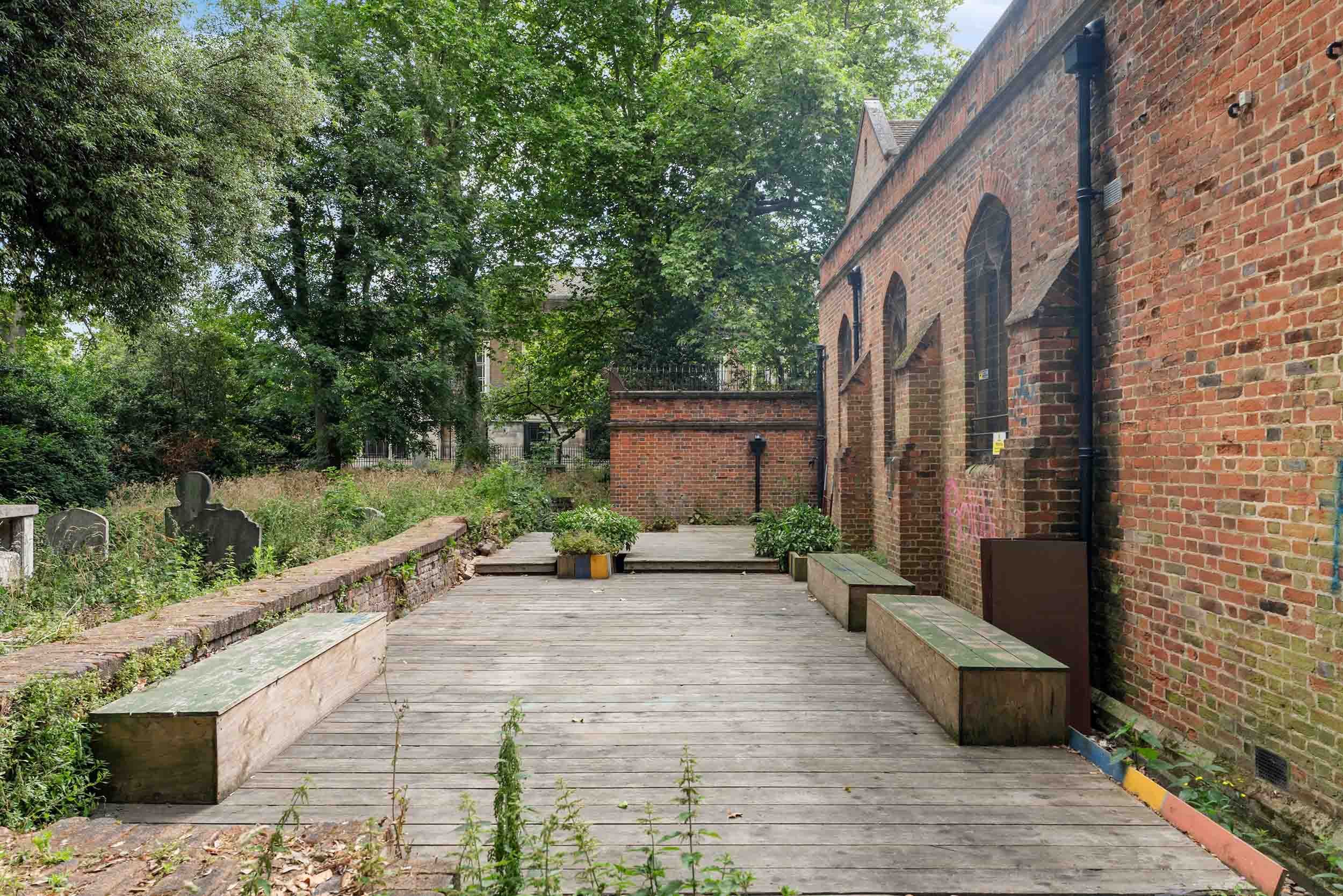 Wooden decking with benches beside a brick building wall, looking out to a grassy churchyard with gravestones.