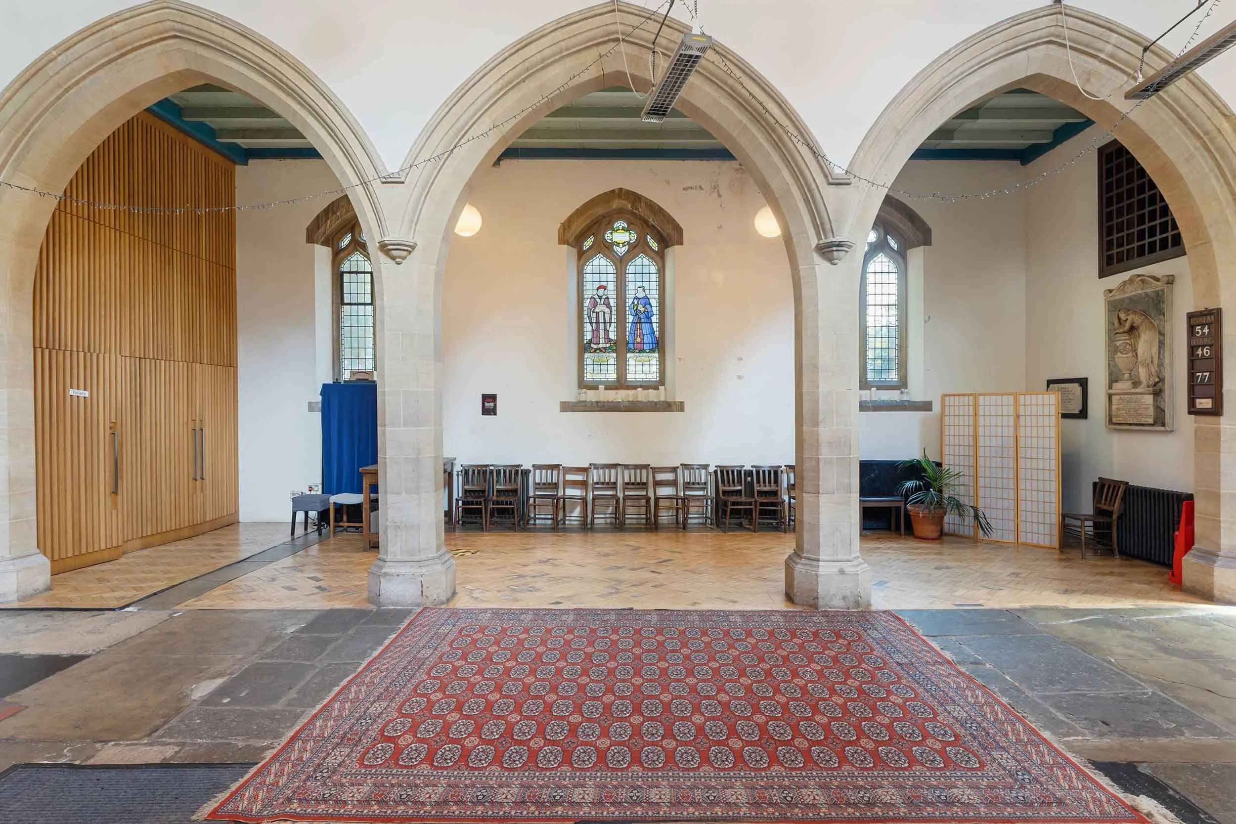 Stone arches framing a herringbone floor and patterned rug, with stained-glass windows and rows of chairs along the wall.