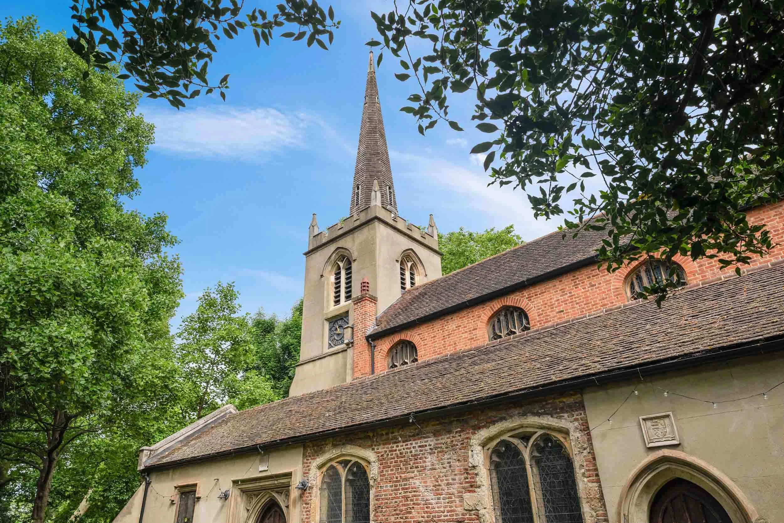 Brick church exterior with arched windows and a tall spire rising above the roofline, framed by trees.
