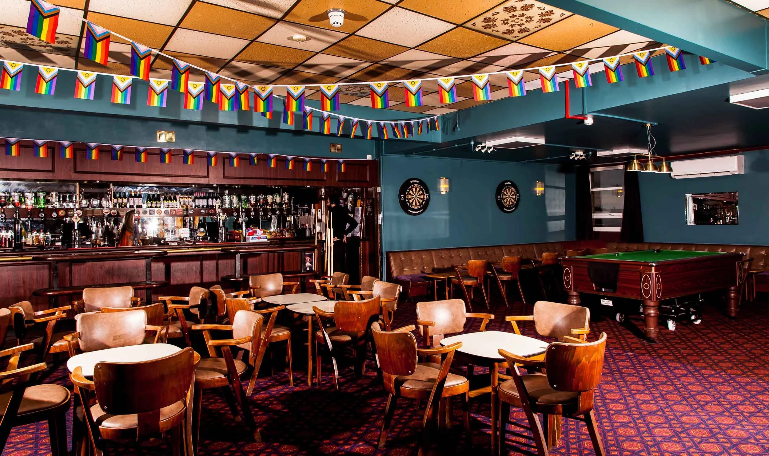 Bar with rows of bottles, wooden tables and chairs, rainbow bunting overhead, and a snooker table beside bench seating.