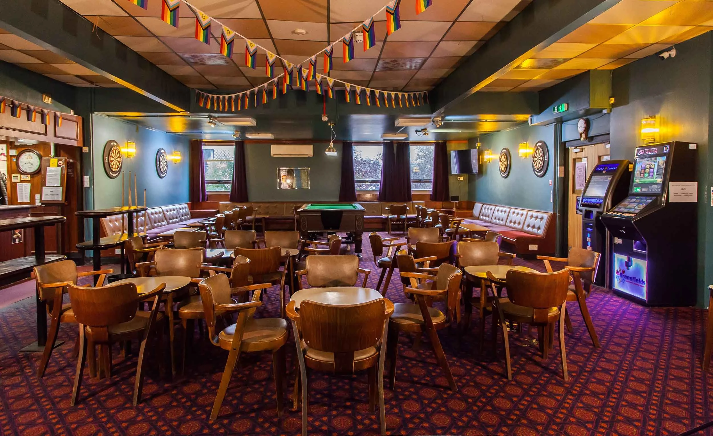 Hall with tables and chairs on patterned carpet, rainbow bunting overhead, dartboards on the walls, a snooker table at the back, and arcade machines to one side.