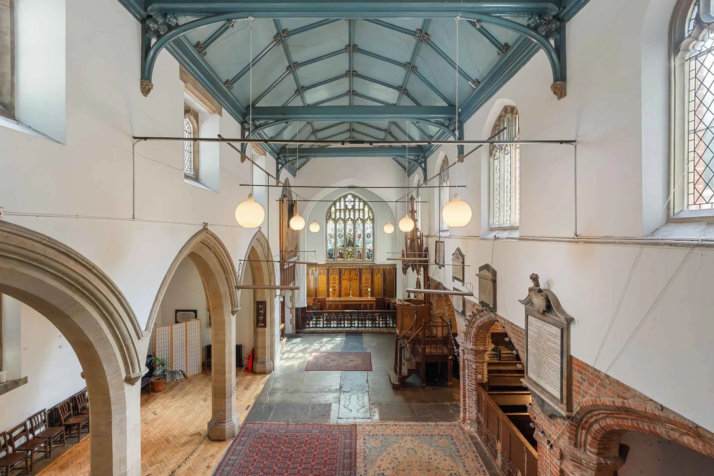 Wide nave with stone arches, rugs on a stone floor, wooden gallery, and hanging globe lights.