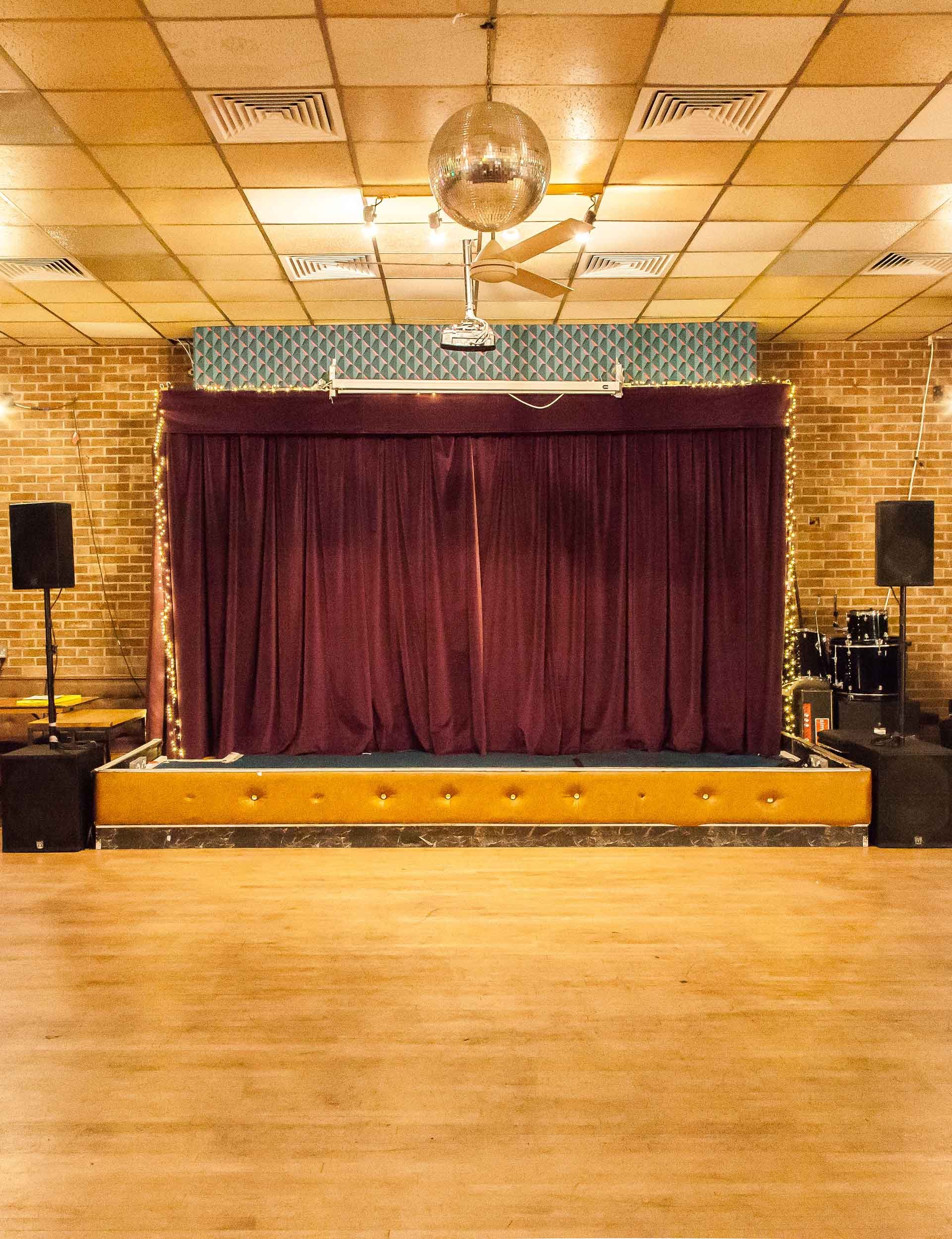 Small stage with closed purple curtain facing a wooden dance floor, with speakers on both sides and a piano to one side.