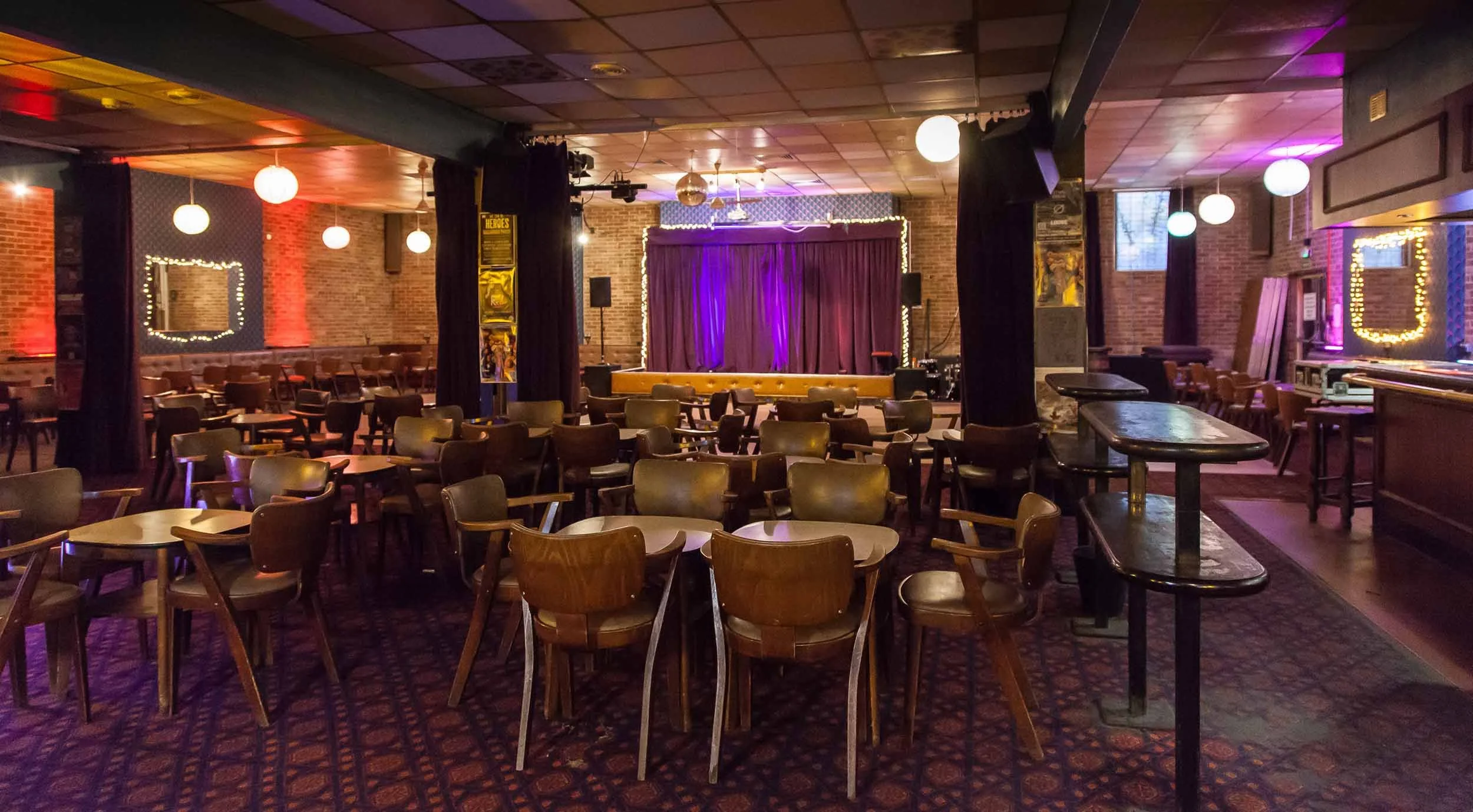 Hall with rows of tables and chairs, patterned carpet, hanging globe lights, and a small stage with purple curtains in the distance.