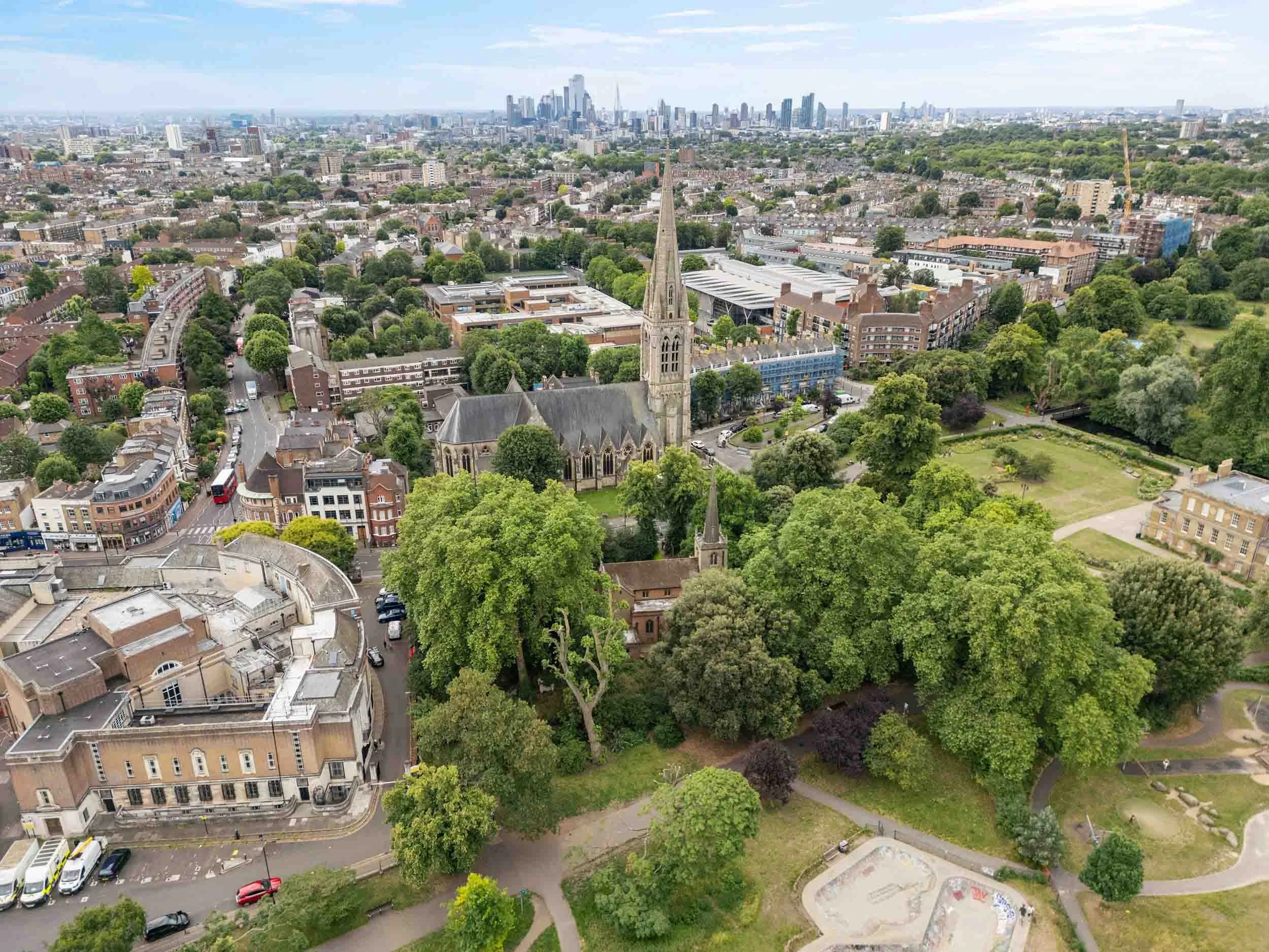 Aerial view of a church with a tall spire beside a park, with the London skyline in the distance.