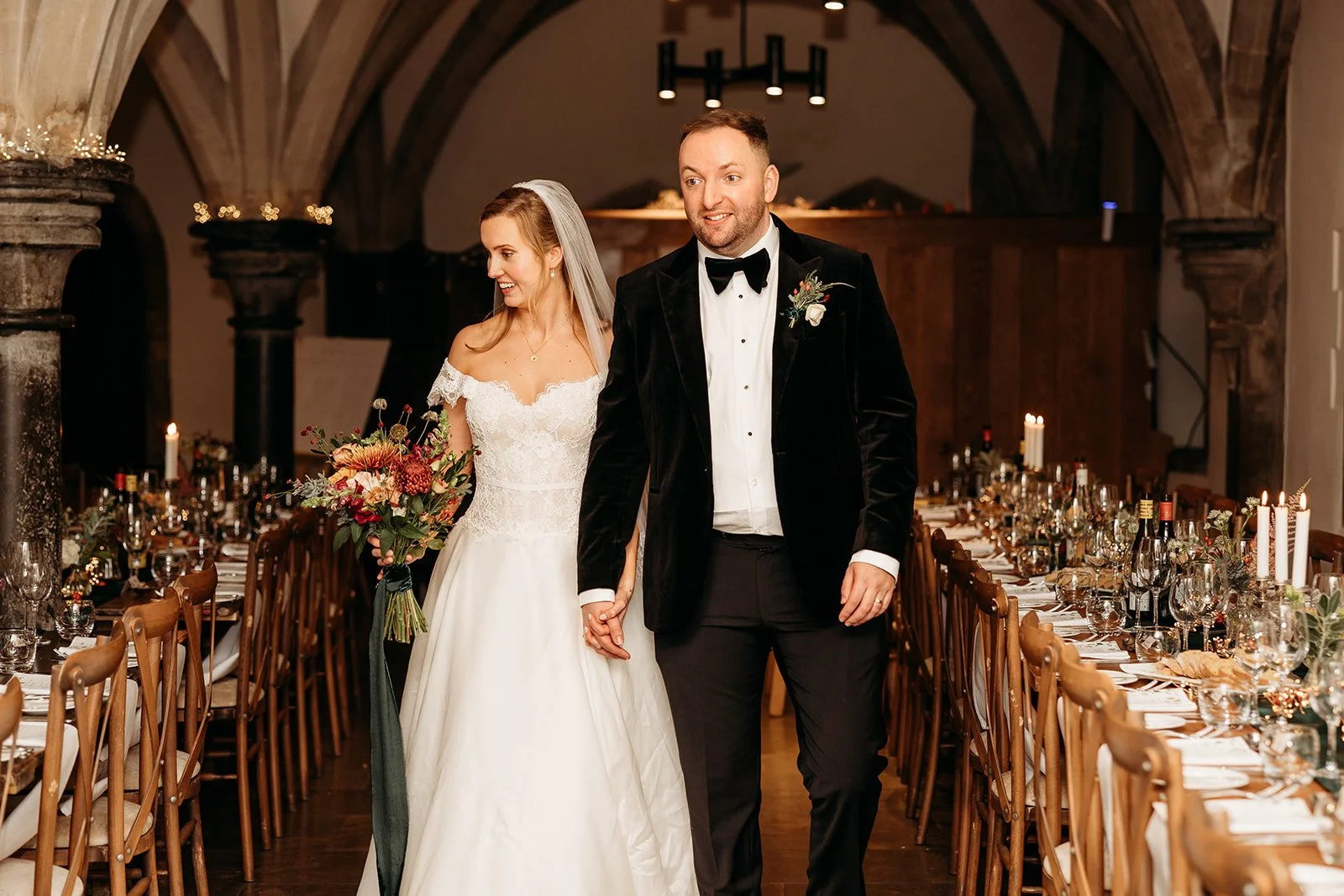 Bride and groom holding hands in a decorated wedding reception hall, with the bride smiling and carrying a large bouquet, banquet tables with candles and floral arrangements in the background.