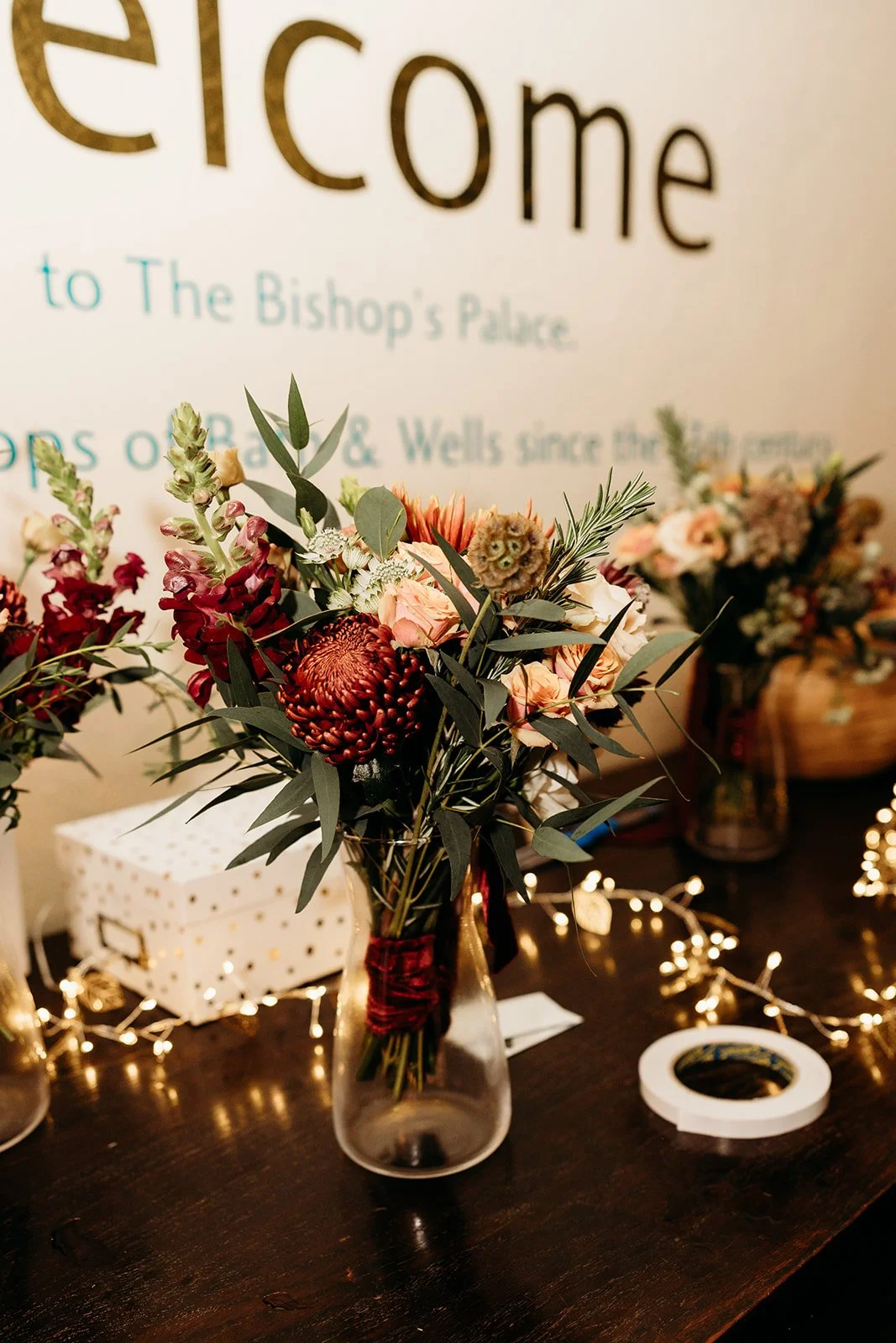 A vase with a floral arrangement of pink and red flowers, greenery, and foliage on a dark wooden table with string lights in the background and a large sign that says "welcome" in the background.