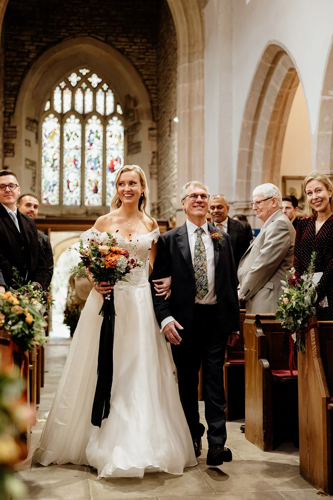 Bride walking down the aisle with her father during a wedding ceremony inside a church with stained glass windows.