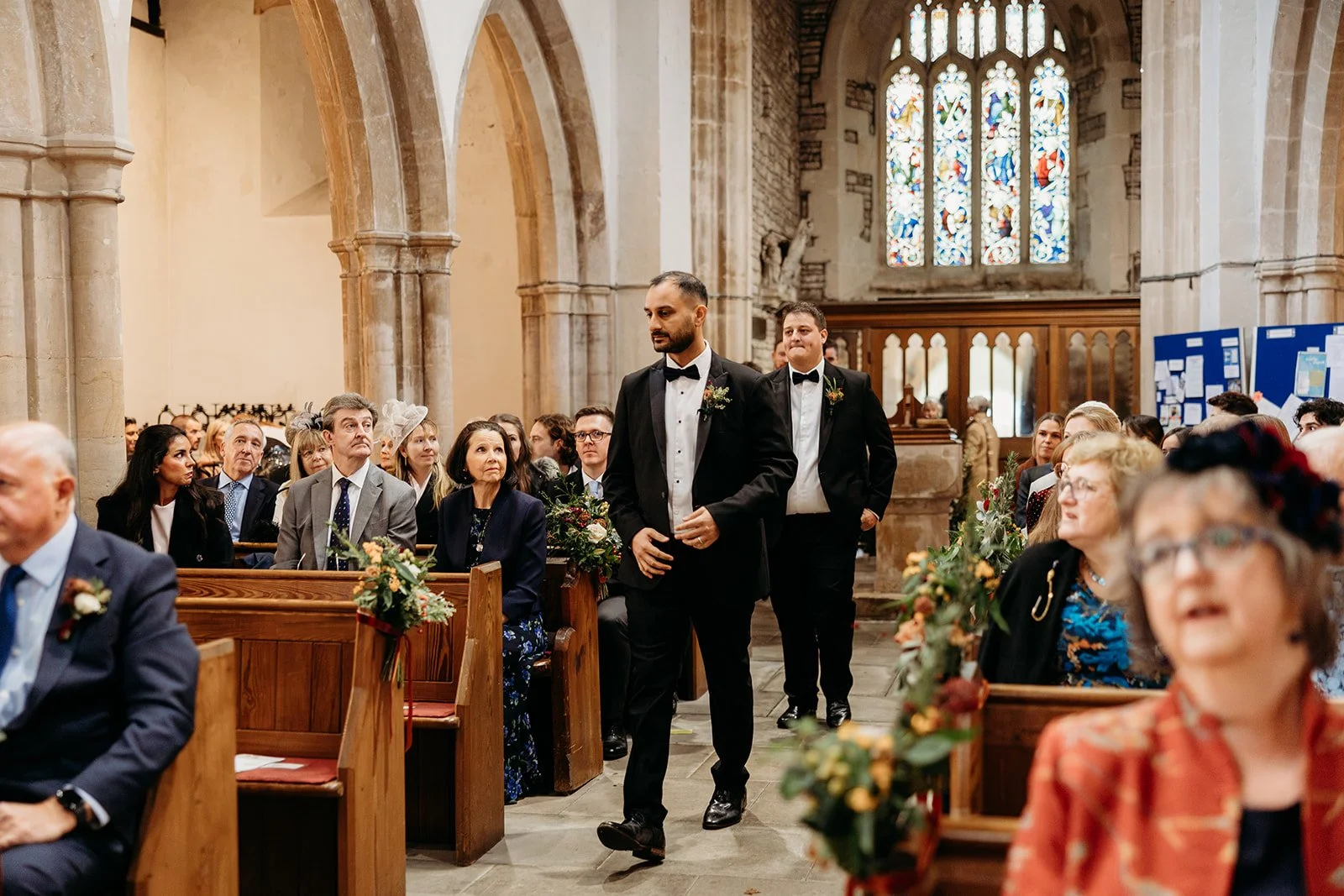A wedding ceremony inside a church with two grooms walking down the aisle, surrounded by seated guests.