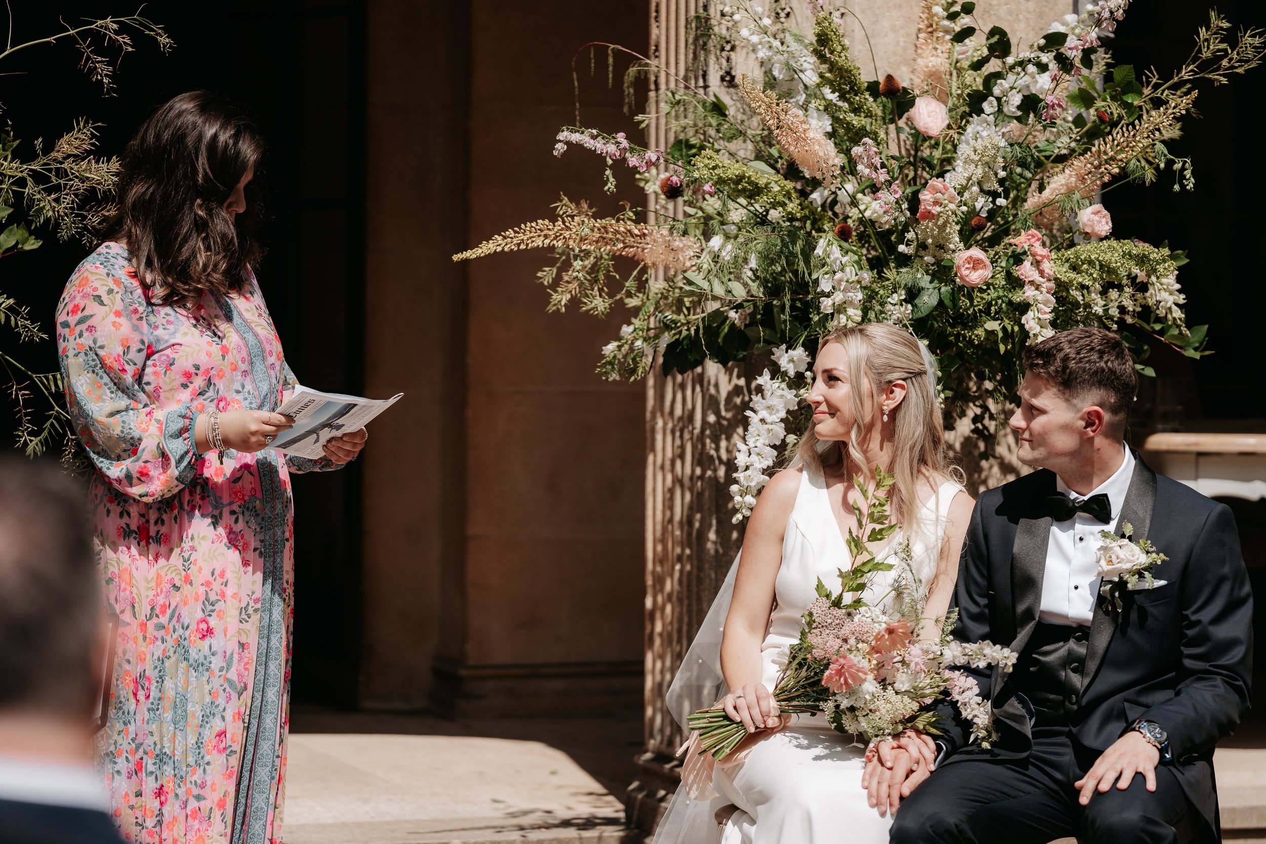 A bride and groom sitting together, holding hands, during their wedding ceremony. The bride is wearing a white wedding dress, and the groom is dressed in a black tuxedo with a bowtie. Both are smiling and looking at the officiant, a woman in a colorf