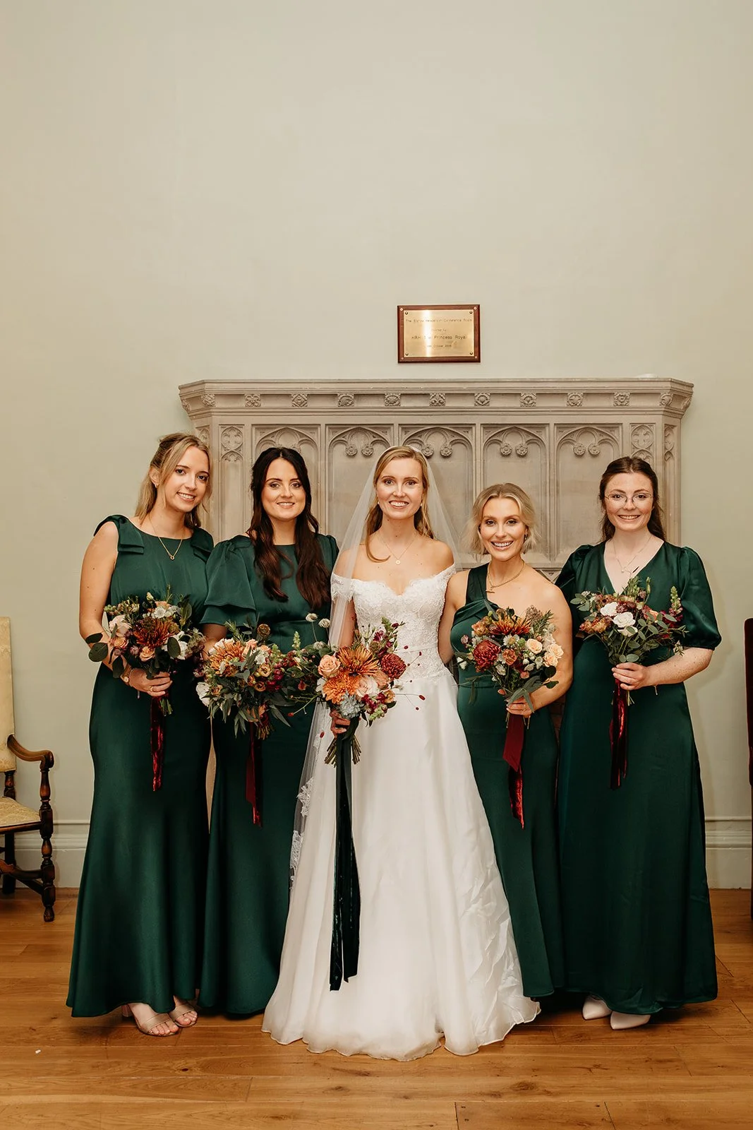 Bride in a white wedding dress with a veil and four bridesmaids in dark green dresses posing in front of a stone fireplace, holding bouquets of flowers.