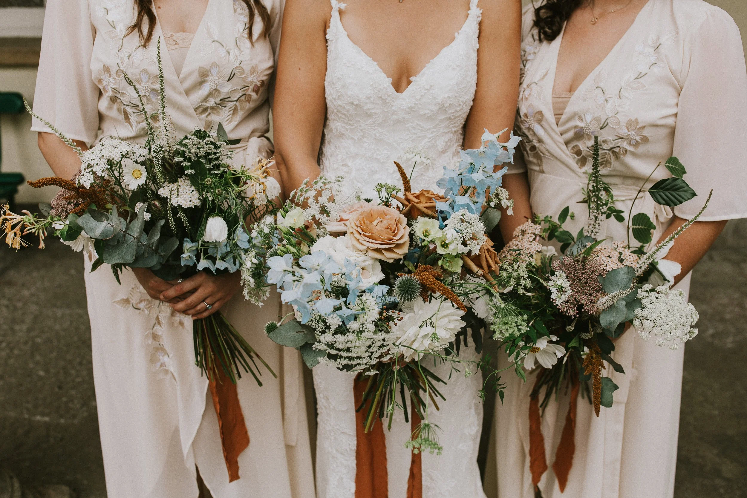Three women dressed in beige and white dresses holding bouquets of mixed flowers and greenery, standing closely together.