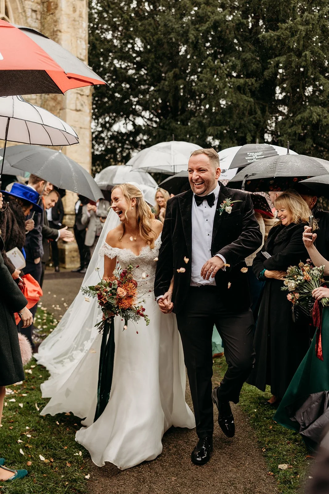 A happy bride and groom holding hands and smiling during their outdoor wedding amid guests holding umbrellas in the rain.